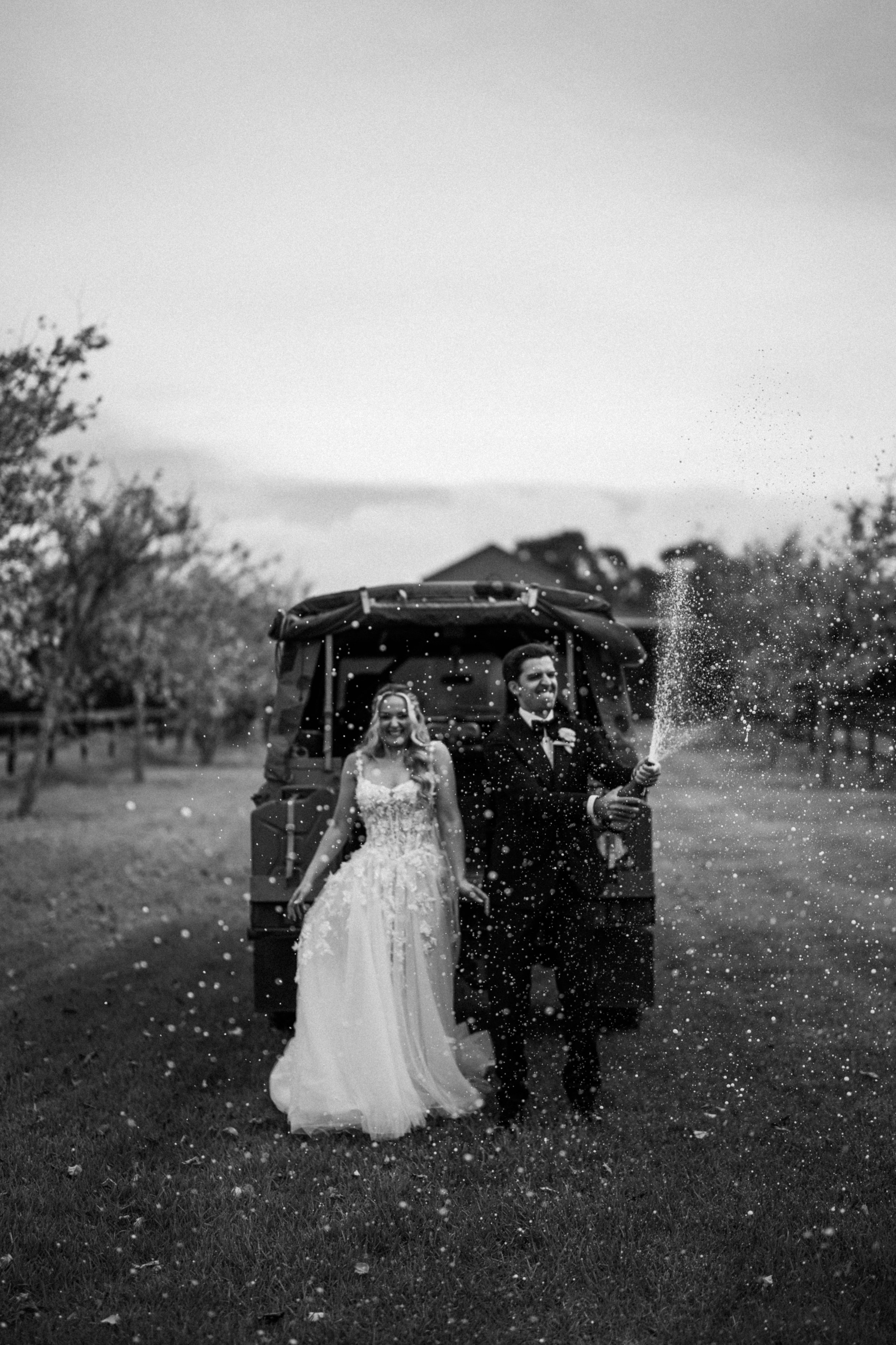 A black-and-white photo of a bride and groom standing outdoors in a grassy area. The groom is on the right, holding a champagne bottle that is spraying. The bride is on the left, smiling, wearing a wedding dress. They are in front of a golf cart, wit