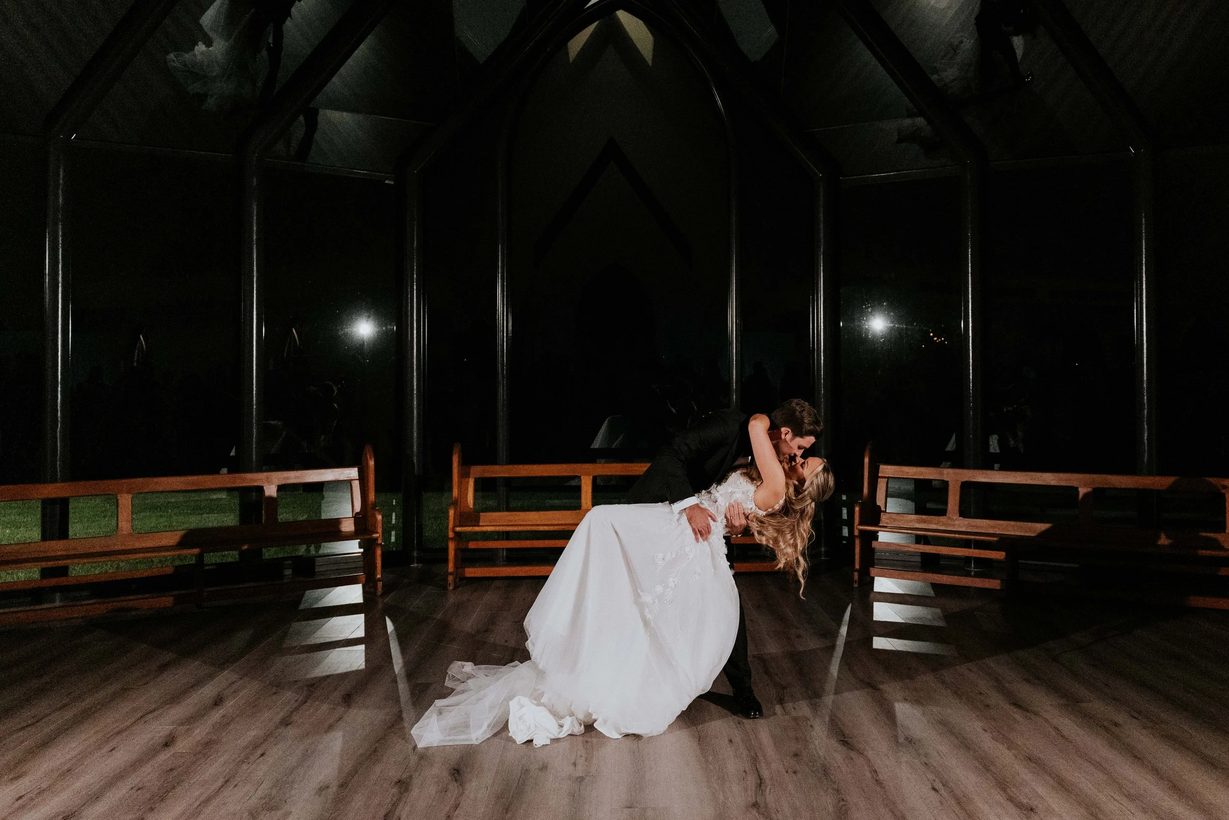 A newlywed couple sharing a romantic dance in a dark, enclosed space with wooden benches and a reflective ceiling, illuminated by soft lighting.