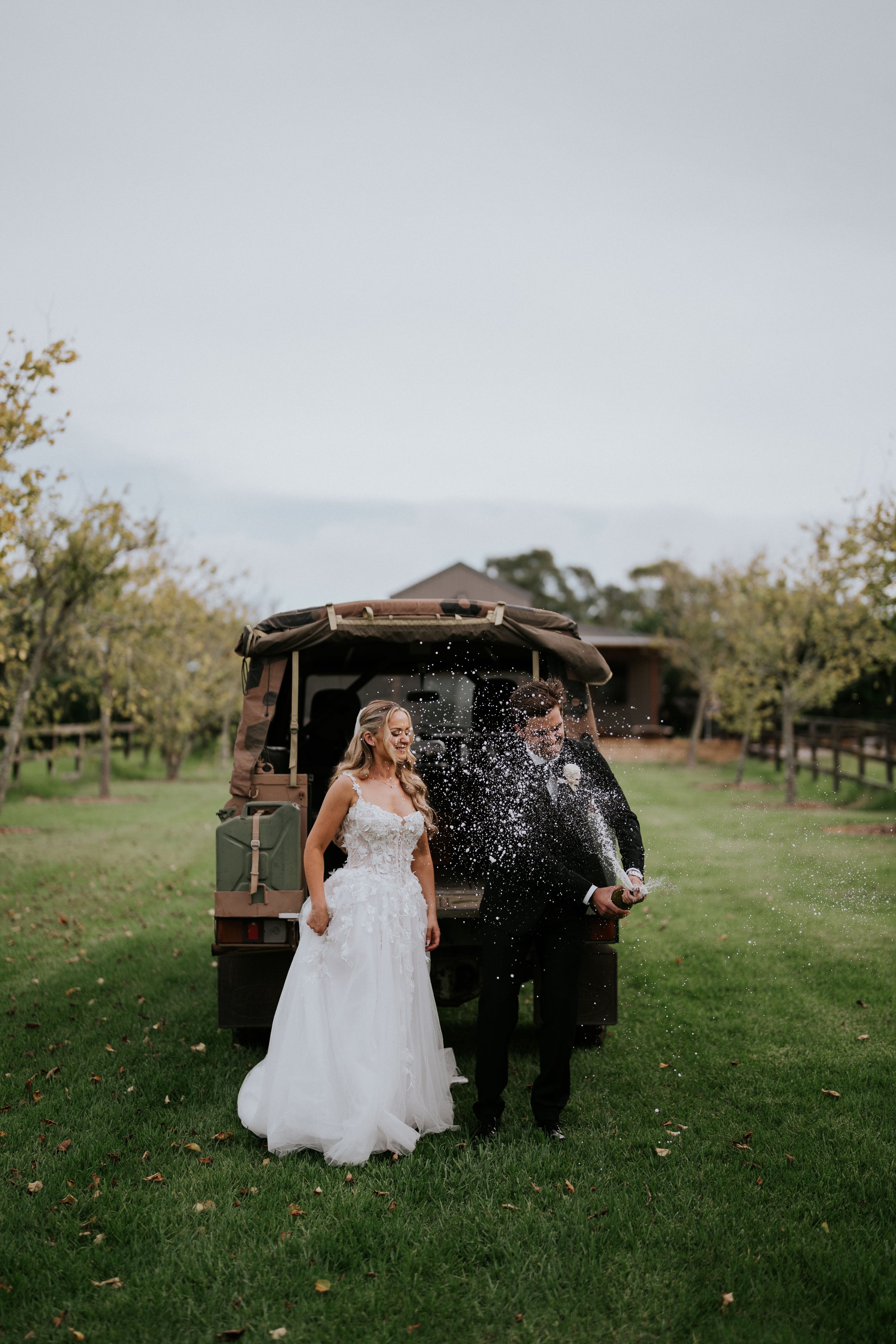 A bride and groom celebrating on a grassy field behind a golf cart, with the groom opening a champagne bottle and spray of champagne in the air, and the bride smiling peacefully.