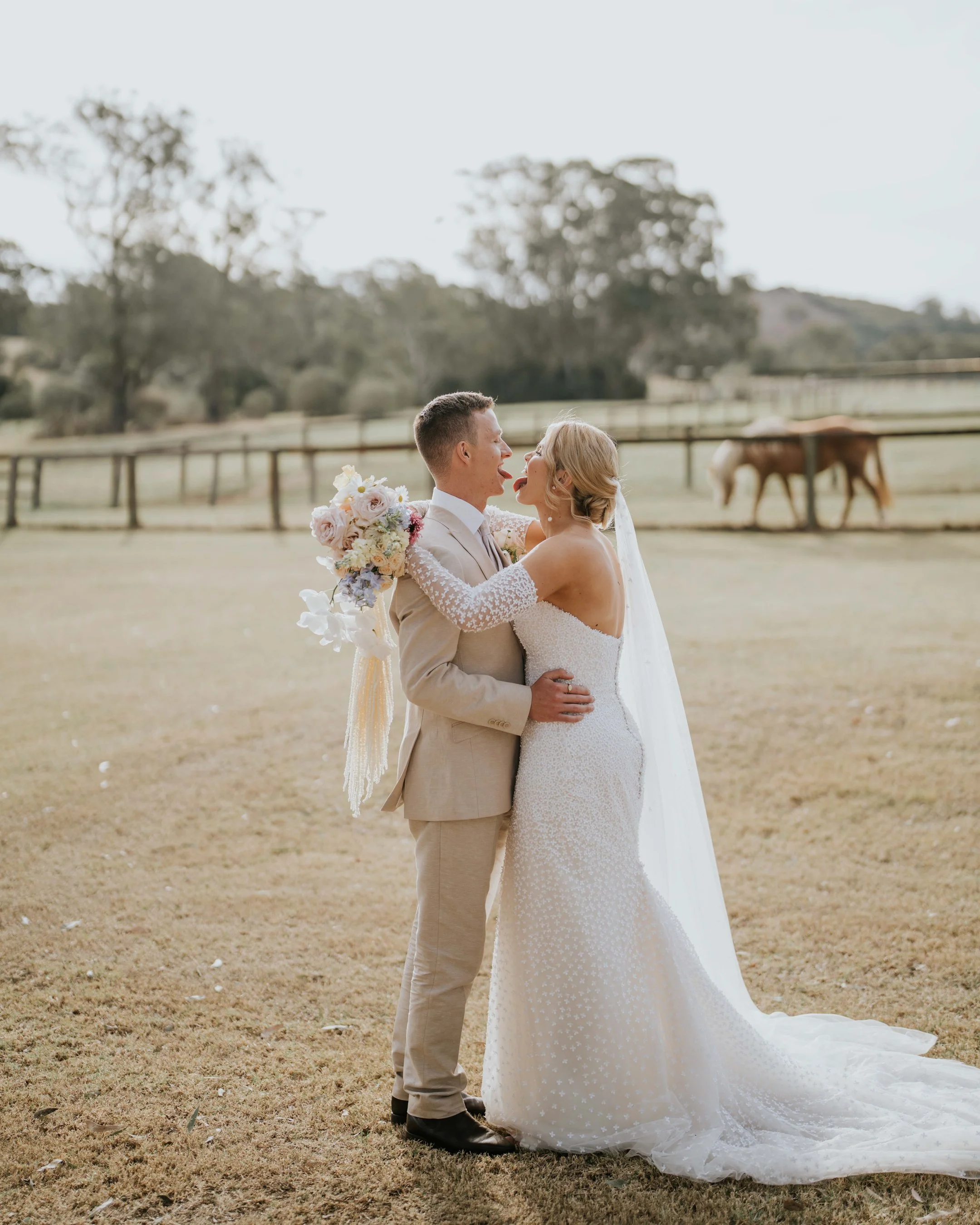 A bride and groom share a playful moment outdoors during their wedding, with the groom sticking his tongue out while the bride touches his face, holding a bouquet of flowers, on a grassy field with trees and a horse in the background.