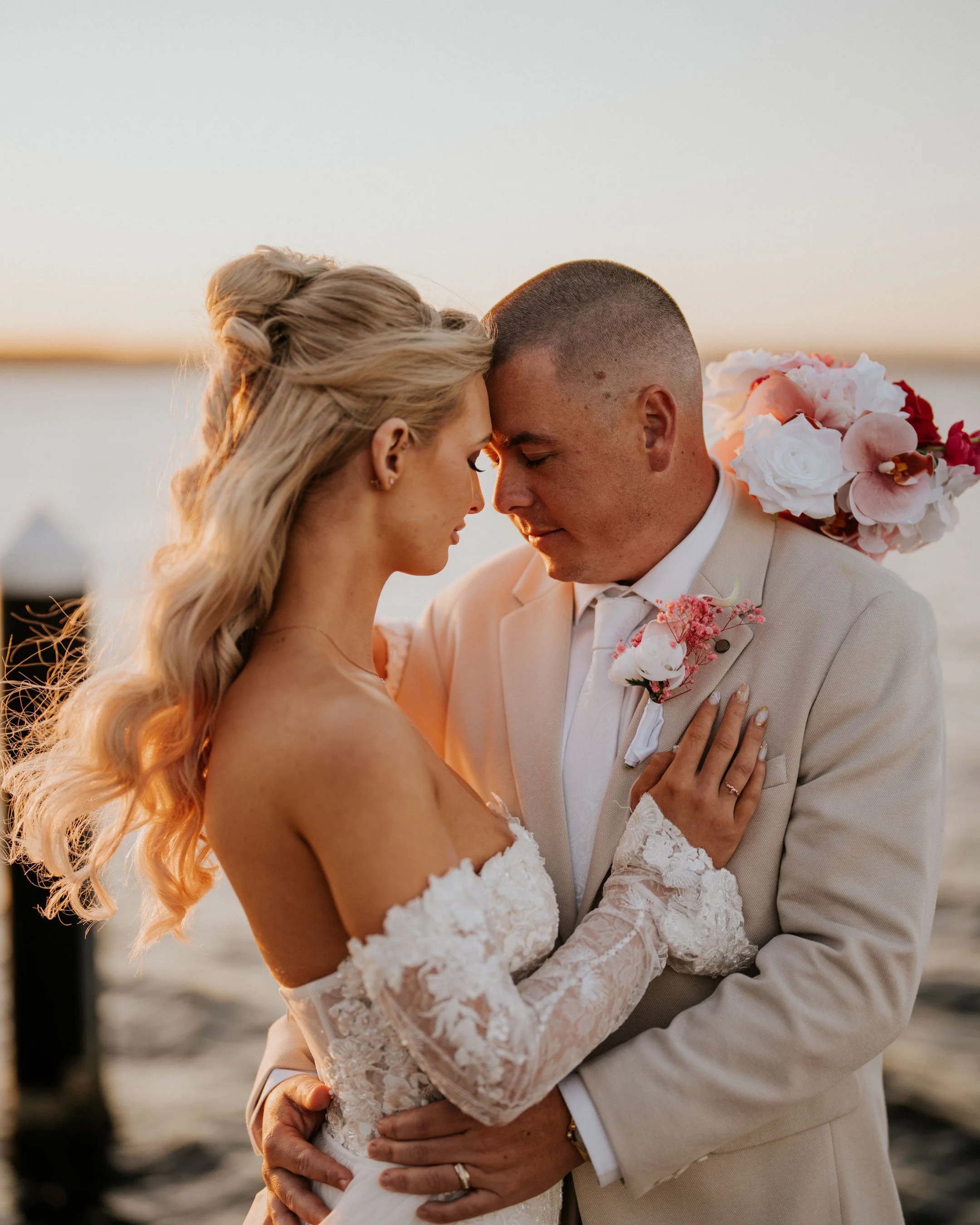 A bride and groom embrace outdoors during sunset, their foreheads touching with eyes closed, in wedding attire with a bouquet of flowers, near water with a sunset sky in the background.