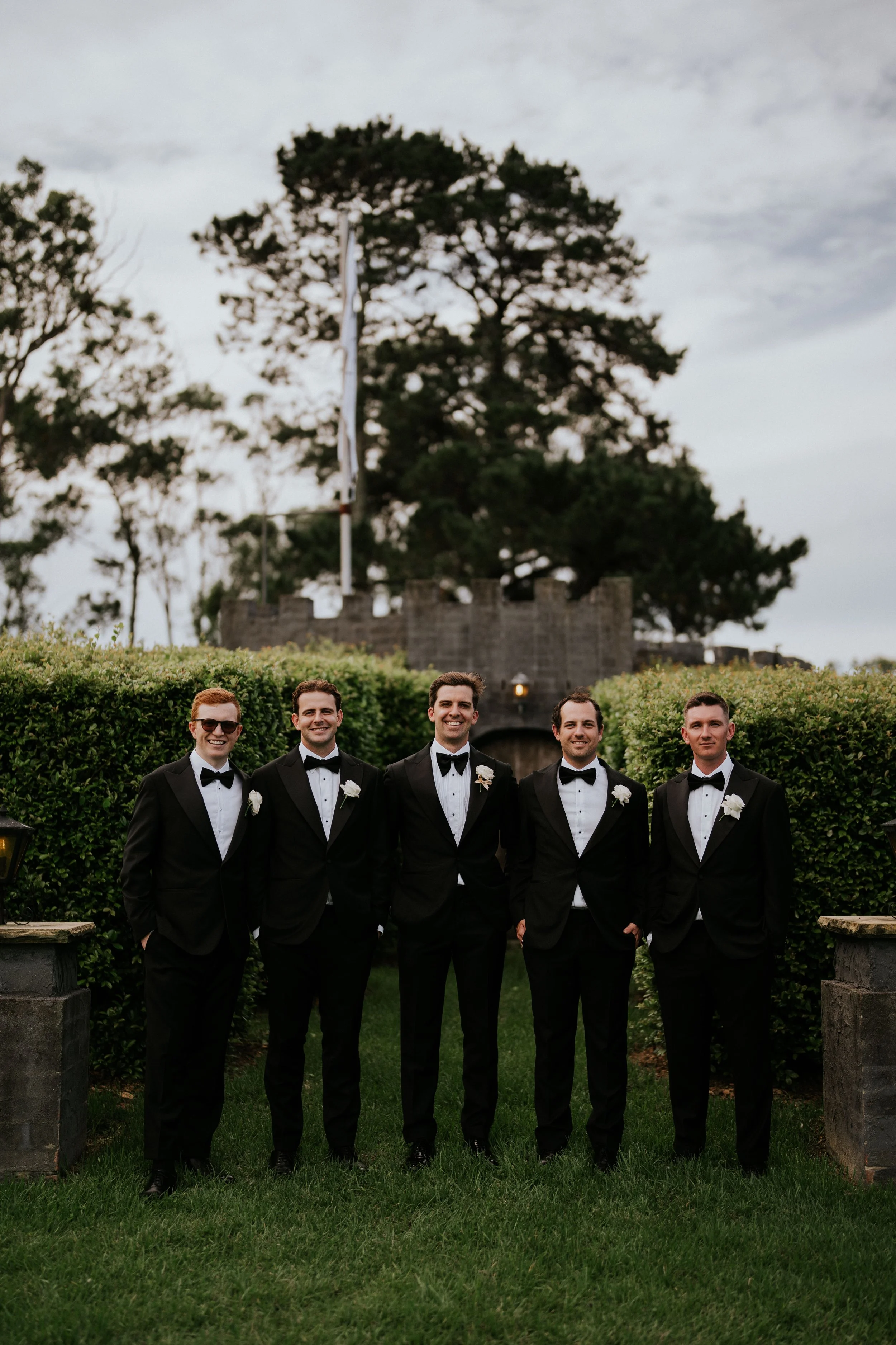 Five men dressed in black tuxedos with white shirts and black bow ties, standing outdoors on grass, smiling. Behind them are green hedges, stone benches, and stone structures with a large tree and cloudy sky in the background.