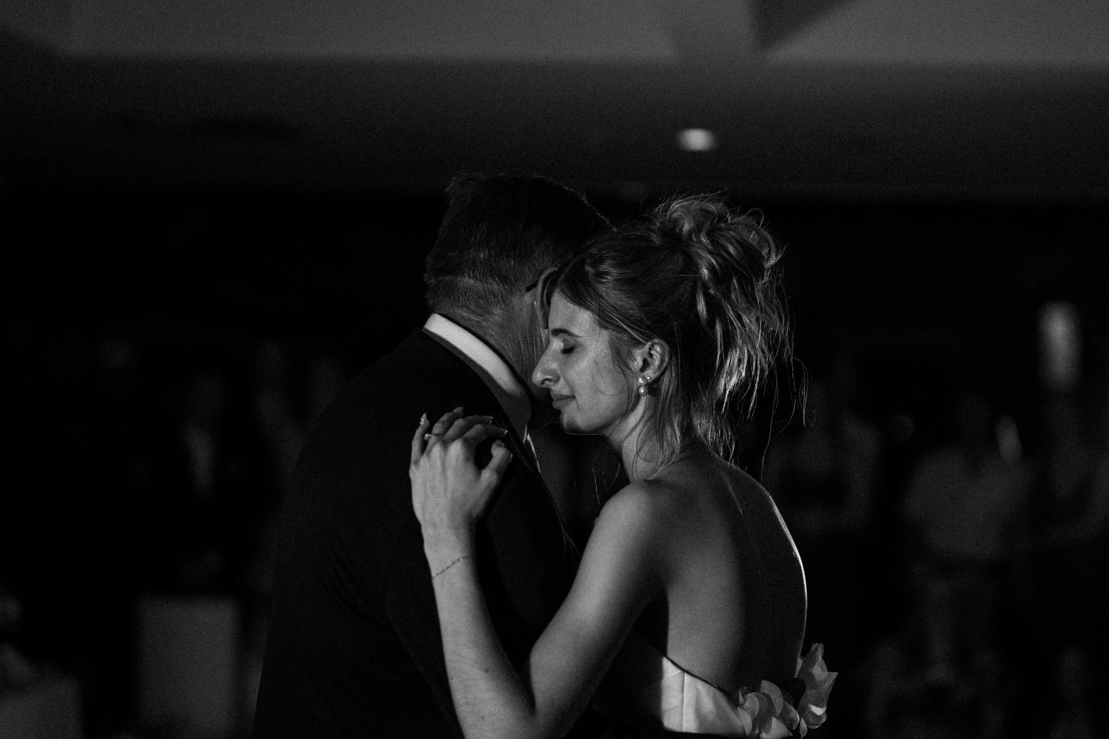 A black and white photo of a couple dancing closely, with the man in a suit and the woman with tousled hair, eyes closed, and a gentle smile, capturing an intimate moment.