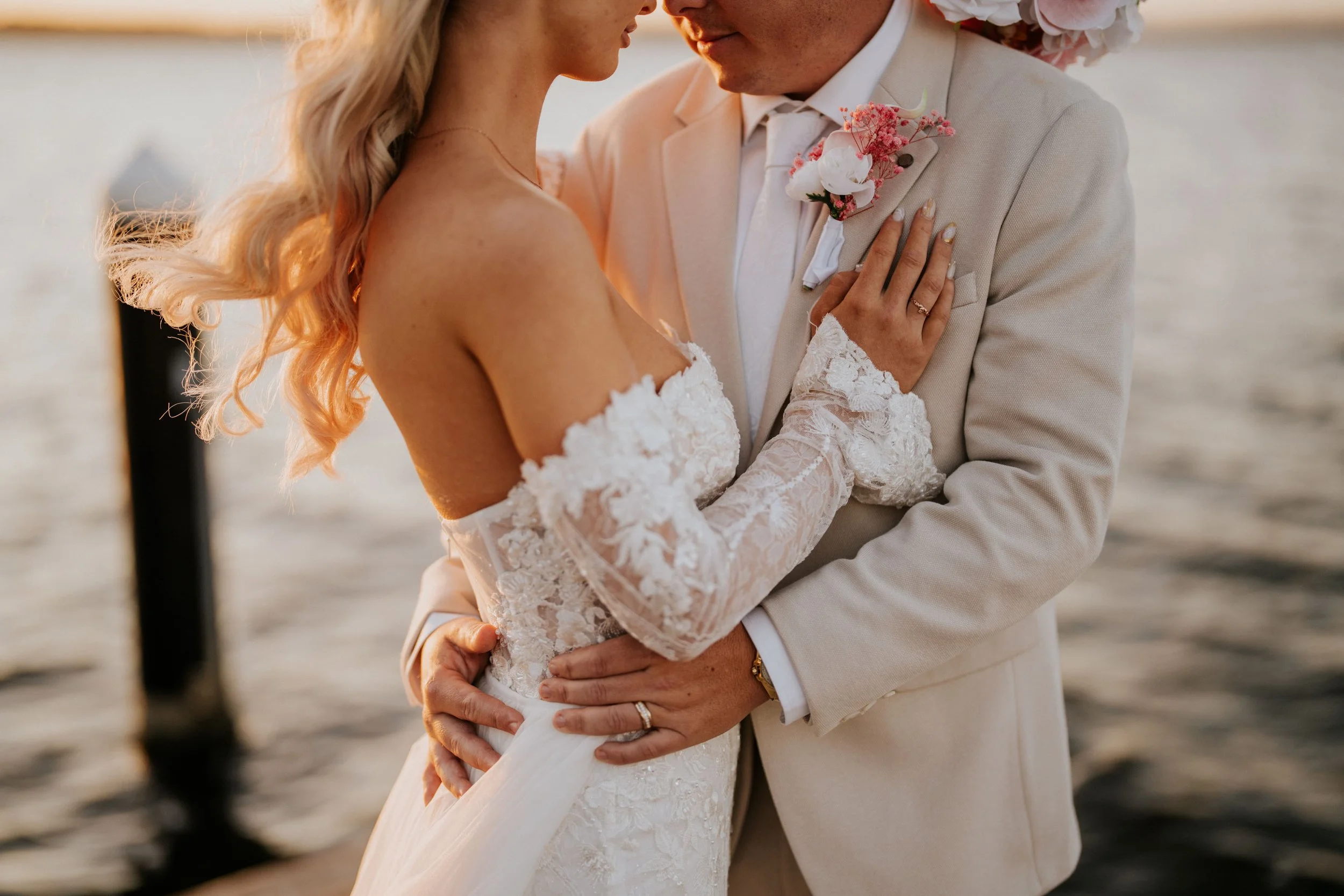 A close-up of a bride and groom embracing outdoors during sunset, focusing on their faces and upper bodies, with the bride wearing a lace off-the-shoulder wedding gown and the groom in a beige suit with a boutonniere, both gently holding each other.