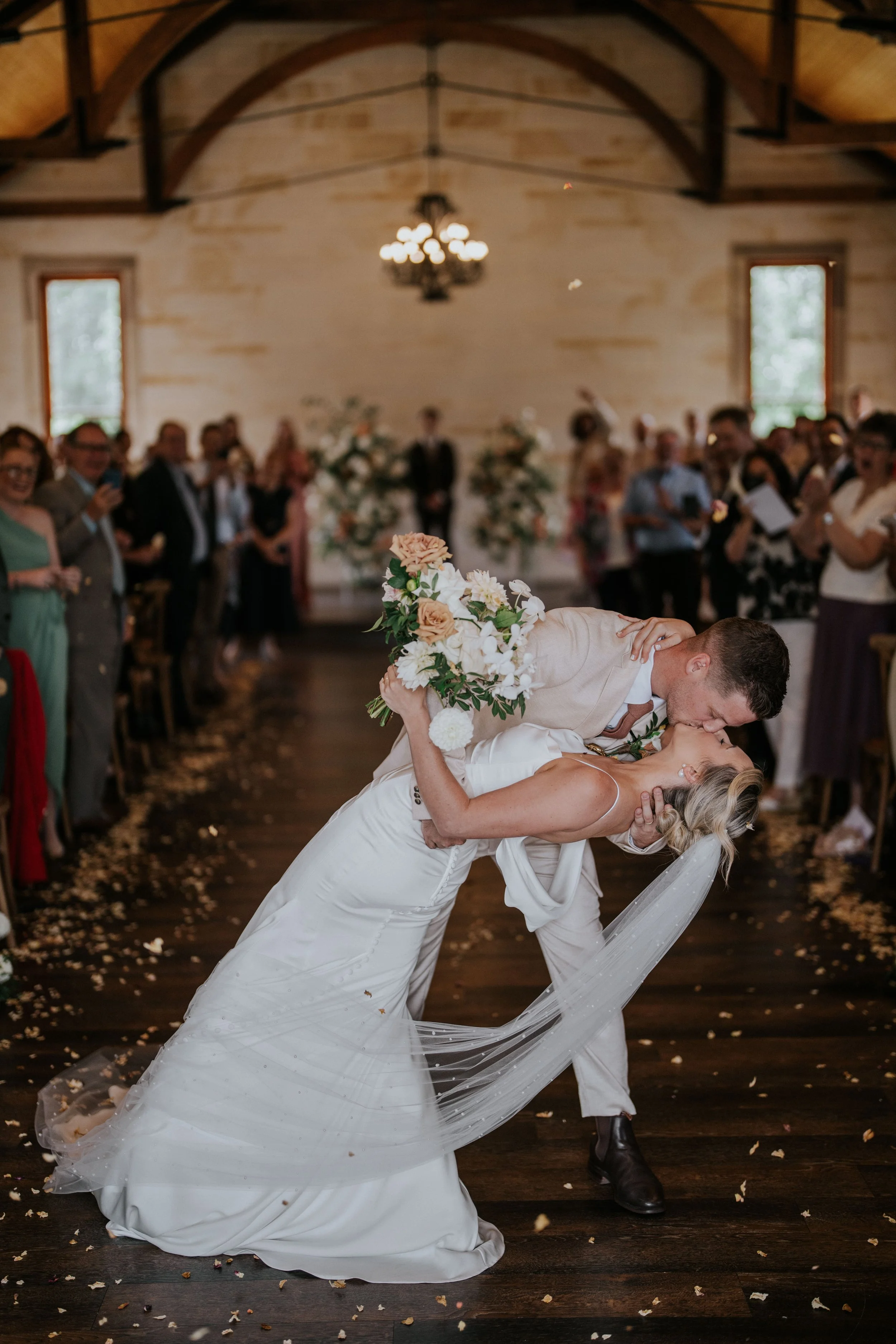A newly married couple dancing at their wedding reception. The groom is dipping the bride and kissing her while holding a bouquet of flowers. The bride’s long white wedding dress has a flowing train. Guests are watching and smiling in the background,