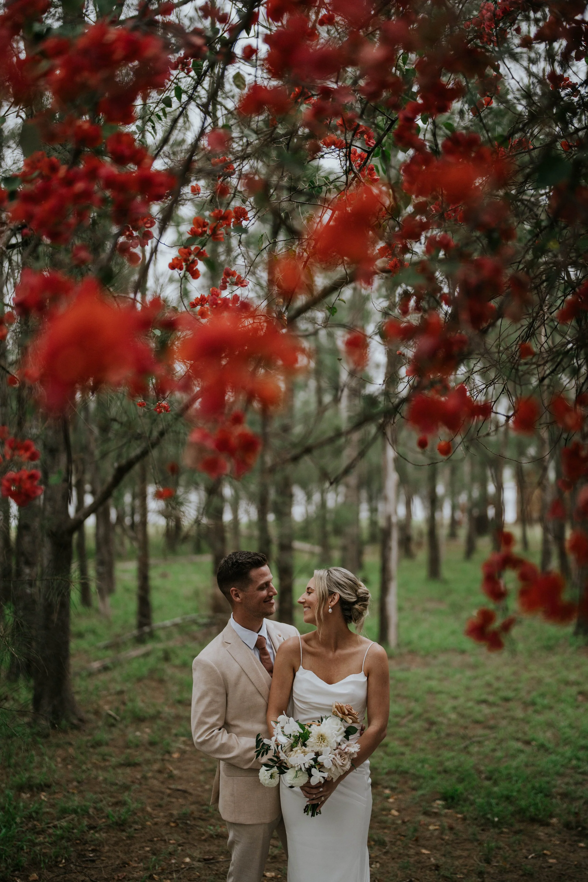 A bride and groom standing outdoors under trees, smiling and gazing into each other's eyes. The bride is holding a bouquet of white flowers, and the groom is wearing a beige suit with a tie. The background features a forested area with tall trees and