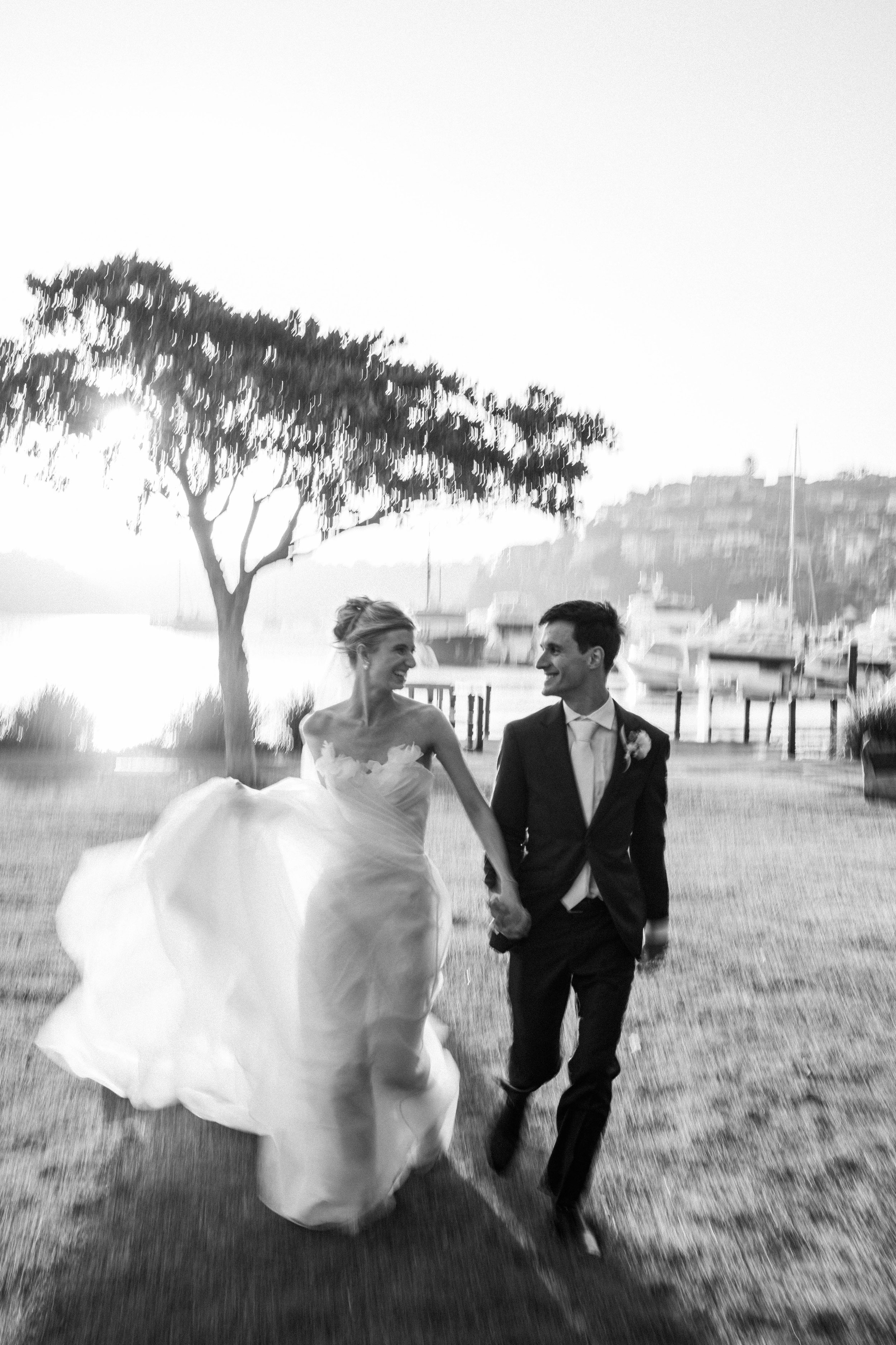 A black and white photo of a bride and groom walking hand in hand outdoors near a body of water. The bride is wearing a wedding dress, and the groom is dressed in a suit with a boutonniere. There is a tree on the left and boats docked in the background.