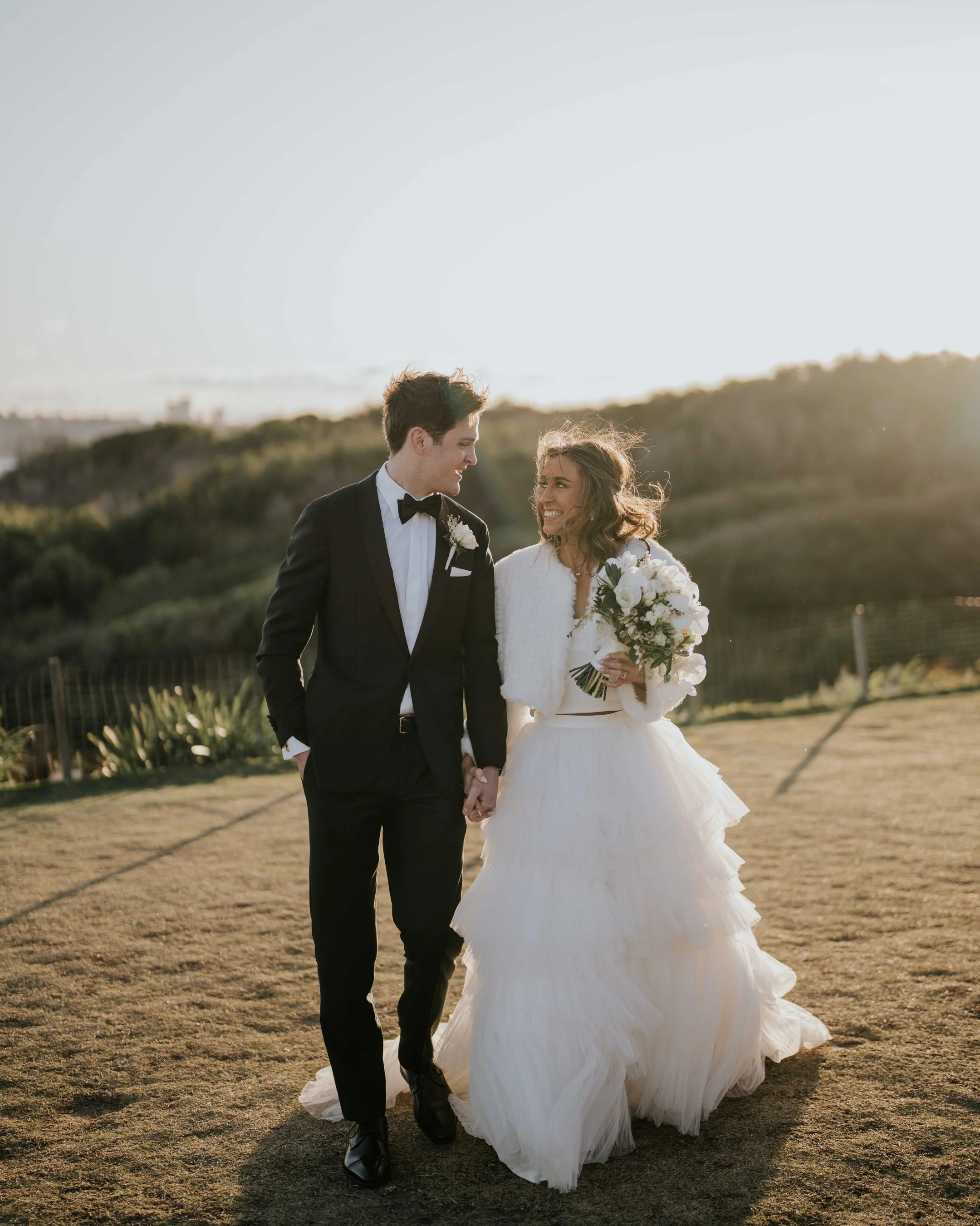 A bride and groom walking hand in hand outdoors, with the sun setting behind them, in a grassy, hilly landscape.
