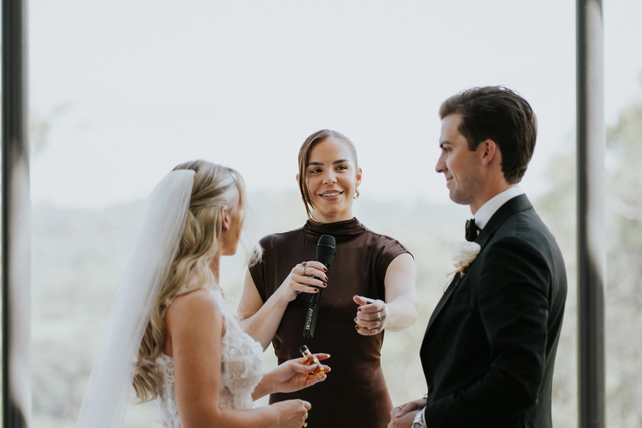 A bride and groom exchanging vows during a wedding ceremony, with a woman officiant holding a microphone and guiding the couple.