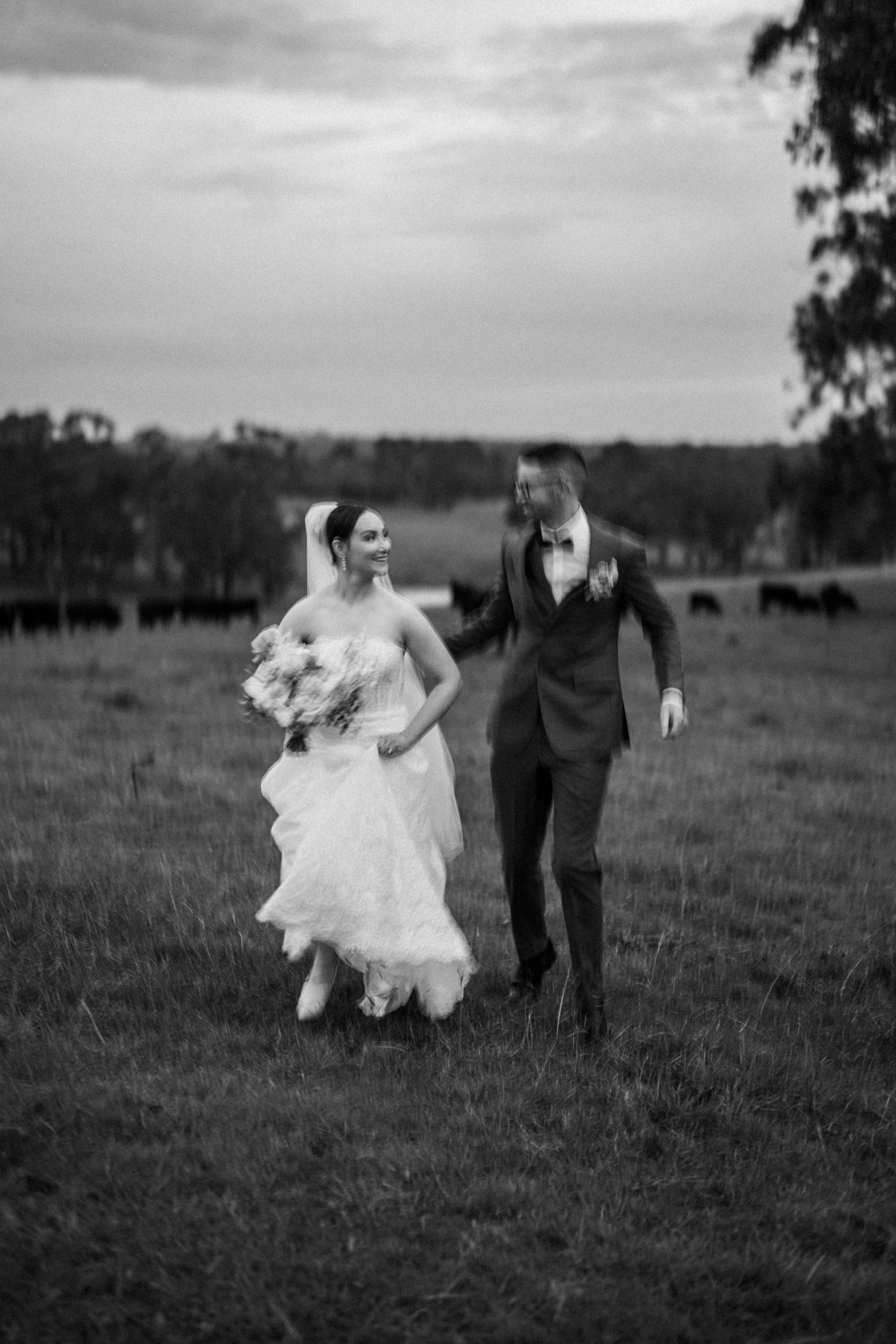 A black-and-white photo of a wedding couple walking through a grassy field, with trees in the background and horses grazing in the distance.