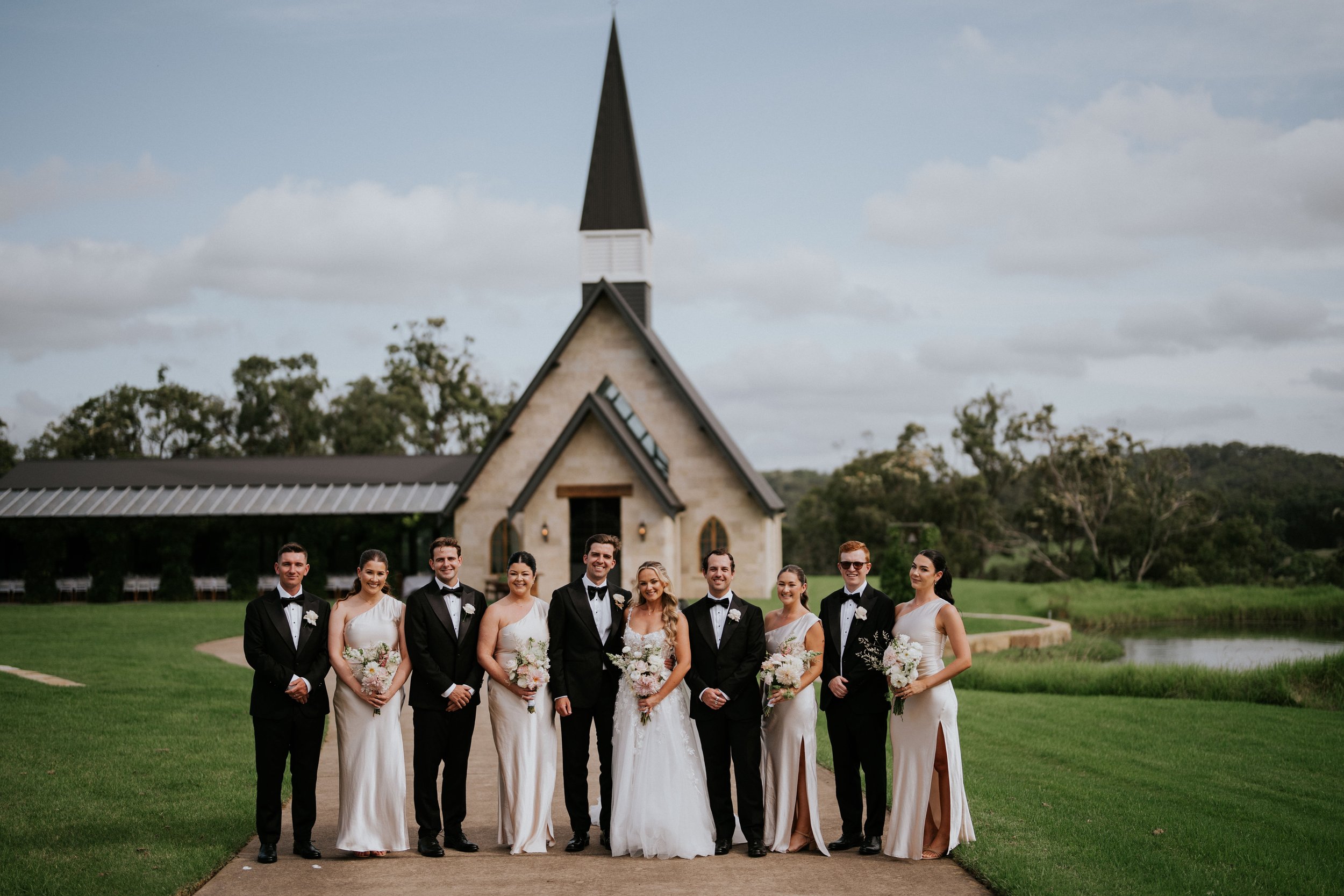 A group of people dressed in formal wedding attire standing outdoors in front of a small church with a tall steeple, surrounded by greenery and trees, under a partly cloudy sky.