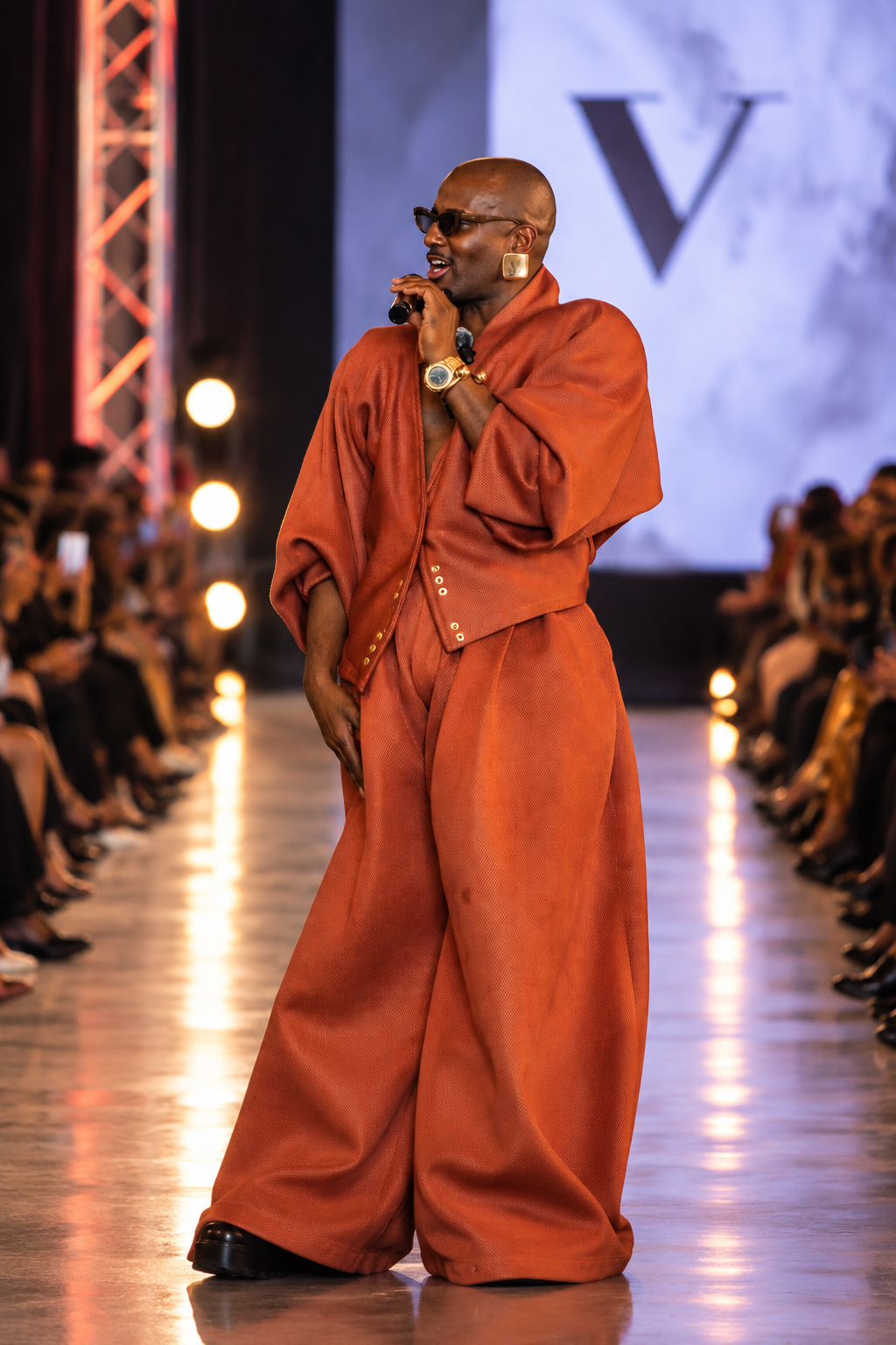 Fashion show presenter in a rust-colored outfit speaking into a microphone on the runway