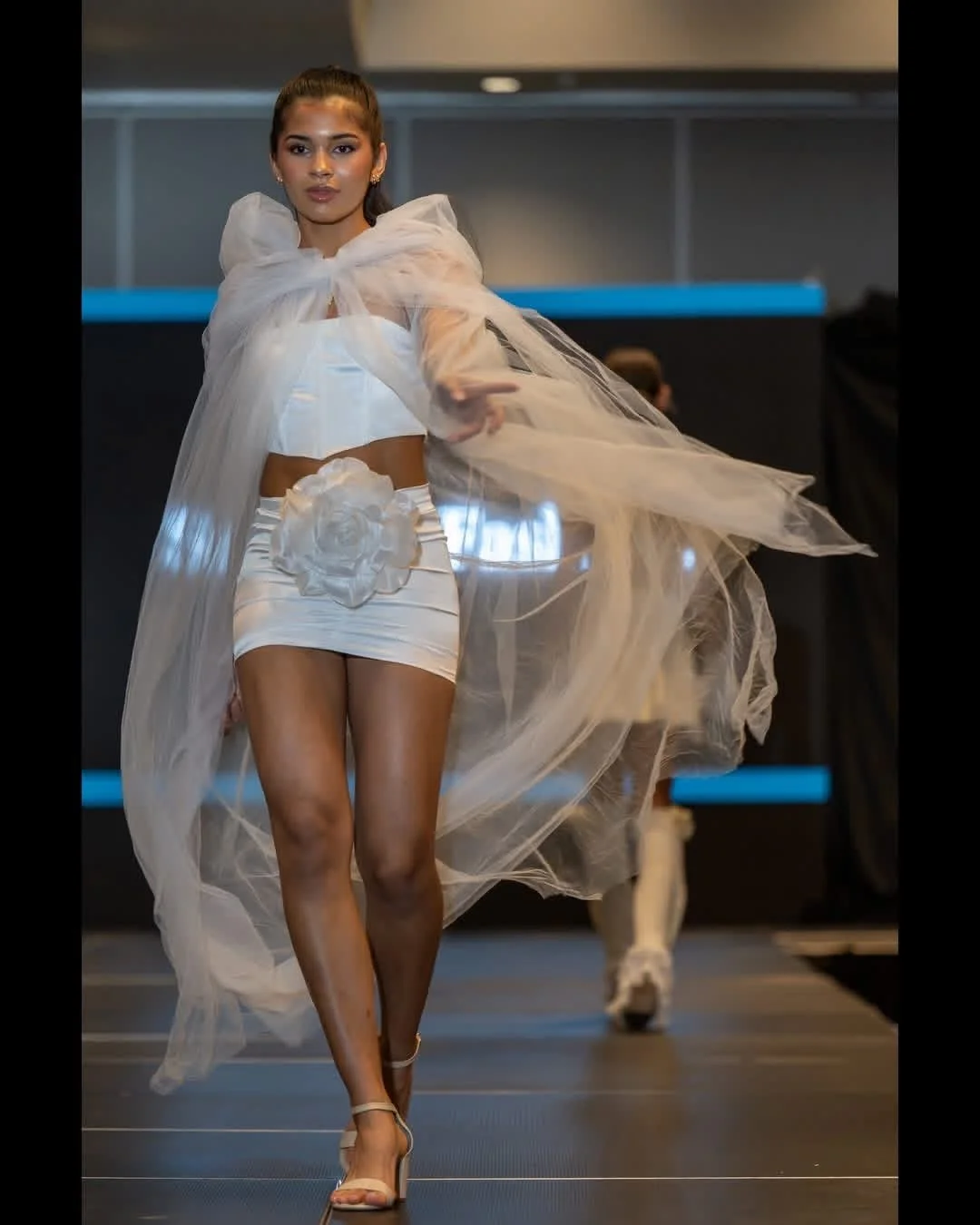 Runway model wearing a white outfit with a flowing sheer cape