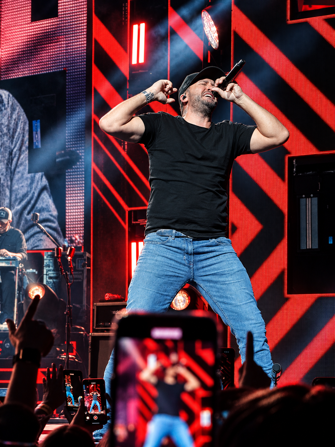 Singer in a black T-shirt performing into a microphone against red stage lights as fans raise their phones