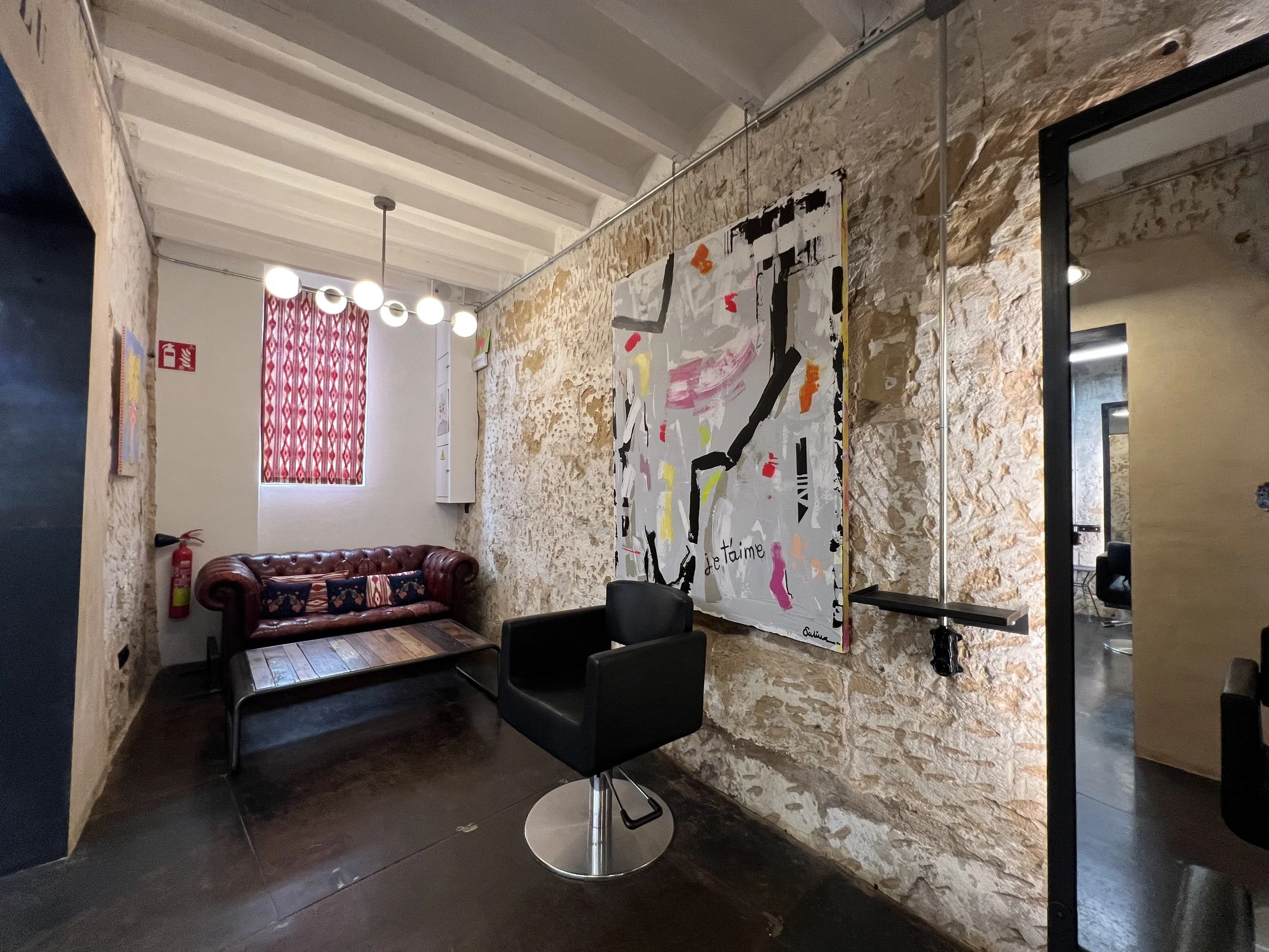 Interior of a room with a textured stone wall, a red patterned curtain over a window, a leather couch with decorative pillows, a fire extinguisher on the wall, and a modern painting with pink and black brushstrokes hanging on the wall. There are black chairs and a small table in the foreground, and a mirror reflecting part of the room.