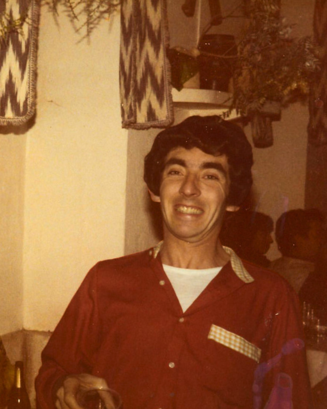 Hombre de cabello oscuro y bigote, sonriendo, con camiseta blanca y camisa roja, sosteniendo un vaso, en un ambiente que parece una reunión o celebración.