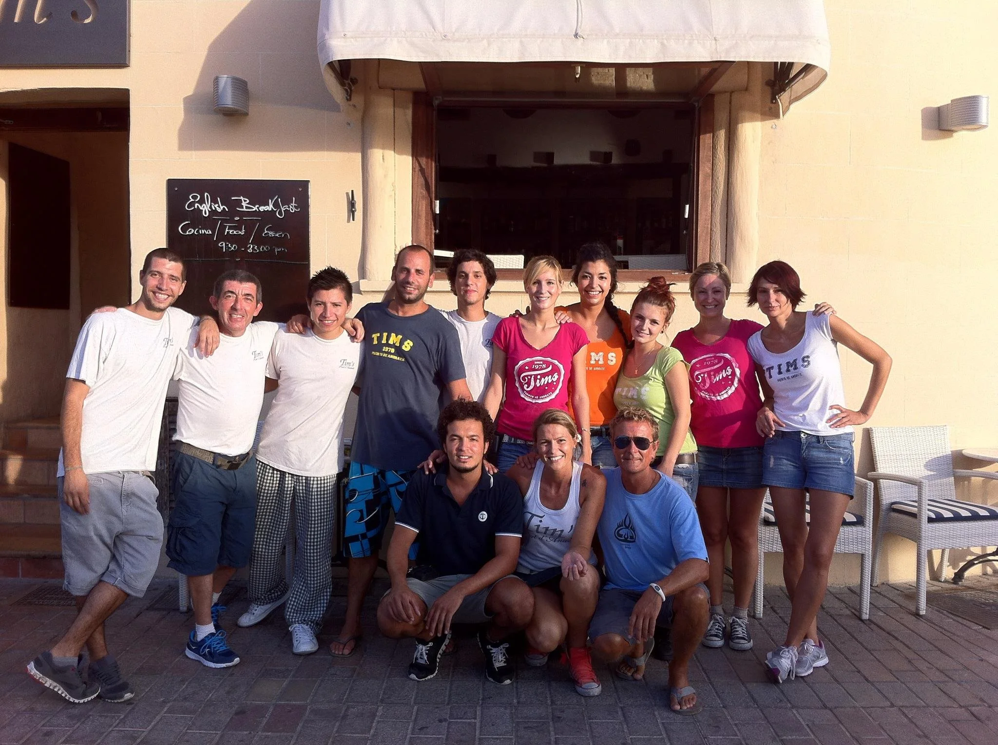 Grupo de personas sonriendo y posando para una foto al aire libre frente a un edificio de color Beige. Se pueden ver algunos carteles y ventanas en el fondo.
