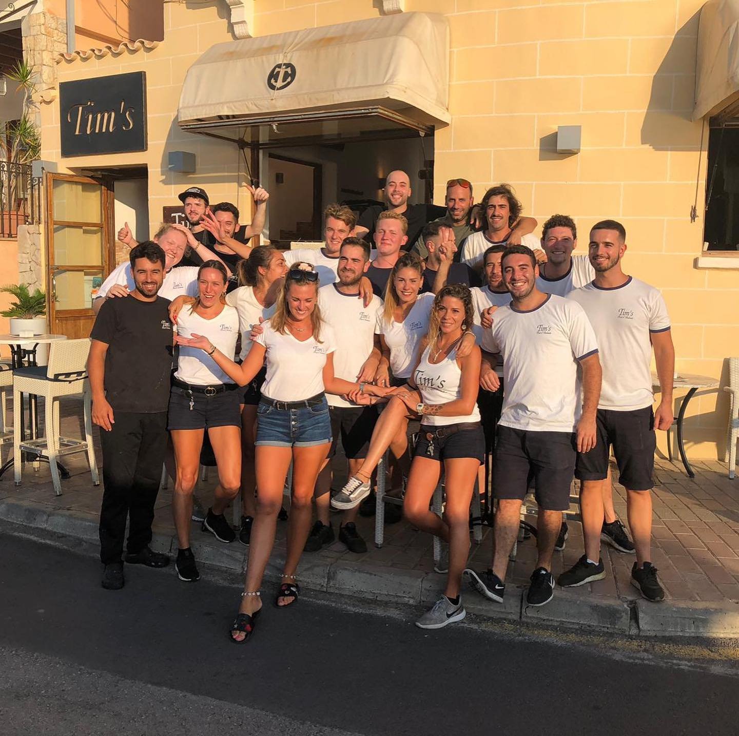Grupo de personas sonriendo y posando frente a un restaurante llamado 'Tim's' al atardecer.