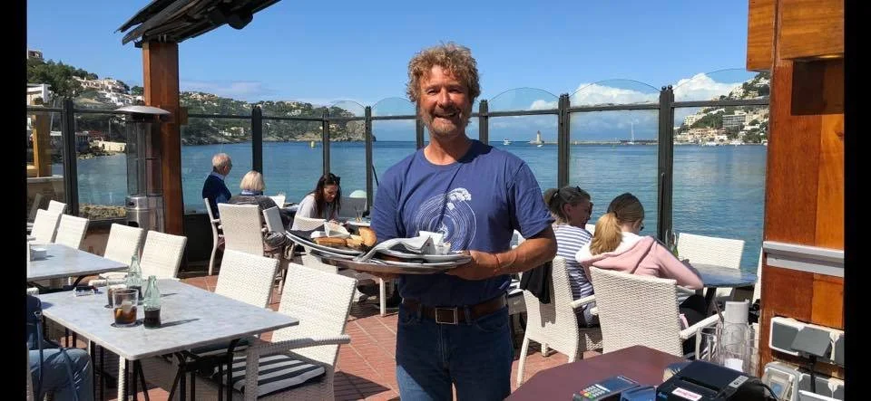 Hombre con barba y cabello rizado sonriendo, sirviendo comida en un restaurante al aire libre con vista a un lago o mar, en un día soleado.