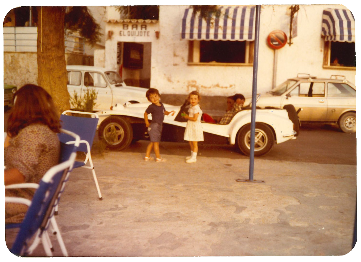 Niños jugando junto a un automóvil deportivo en la calle, con adultos sentados en sillas y unos edificios con toldos a rayas en el fondo.