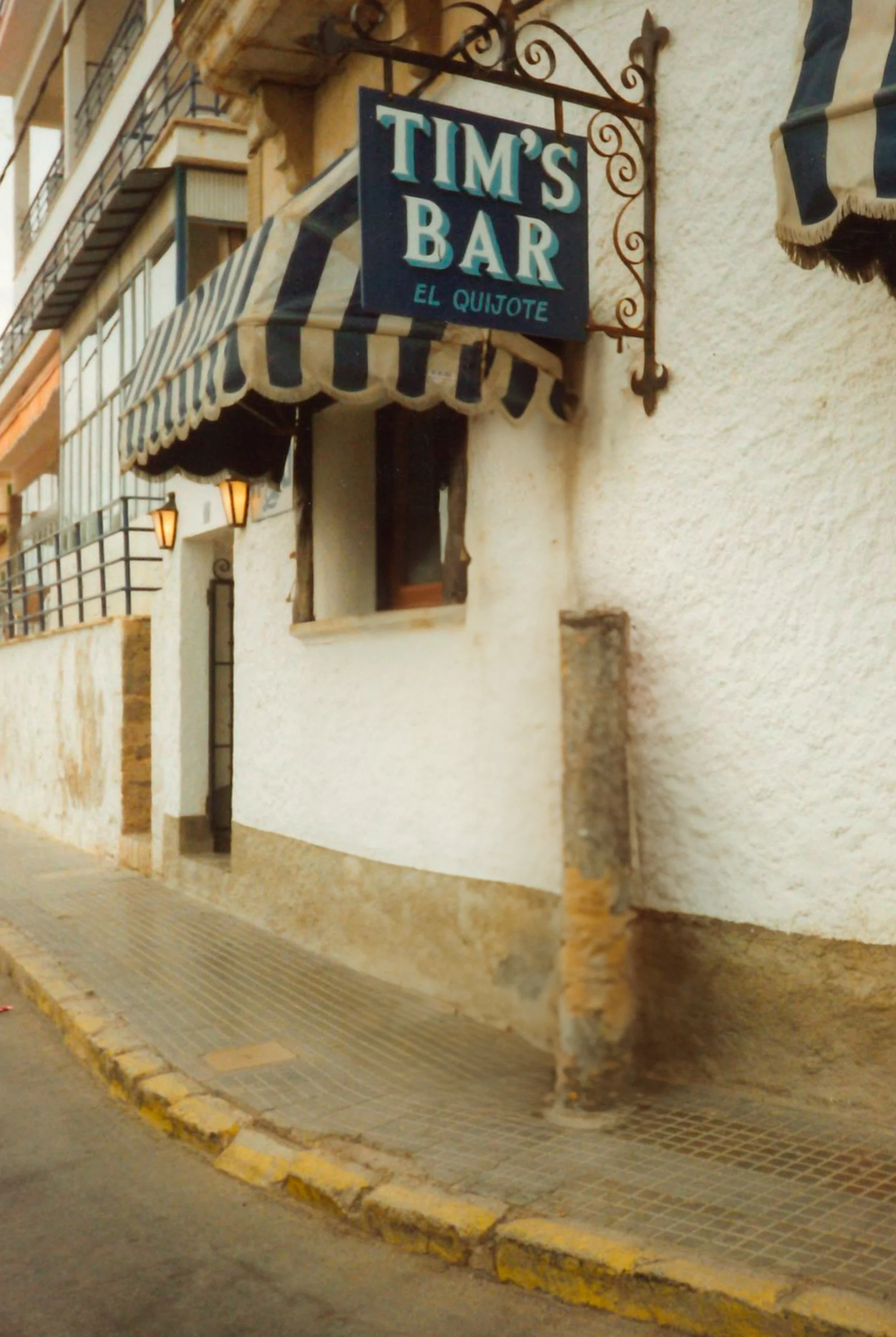 Cartel de un bar llamado 'Tim's Bar El Quijote' en una pared de un edificio con una marquesina a rayas y lámparas en la fachada.