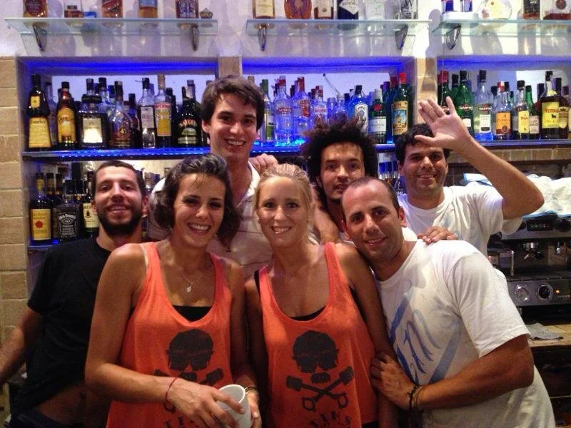 Grupo de seis personas en un bar, sonriendo y posando para la foto. Detrás, estanterías con muchas botellas de alcohol. Algunos llevan camisetas naranjas con un diseño de calavera y tijeras.