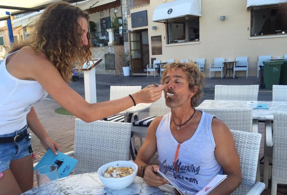 Una mujer le está dando de comer a un hombre con una cuchara en una mesa de un restaurante al aire libre, en un entorno urbano con sillas blancas y edificio de fondo.