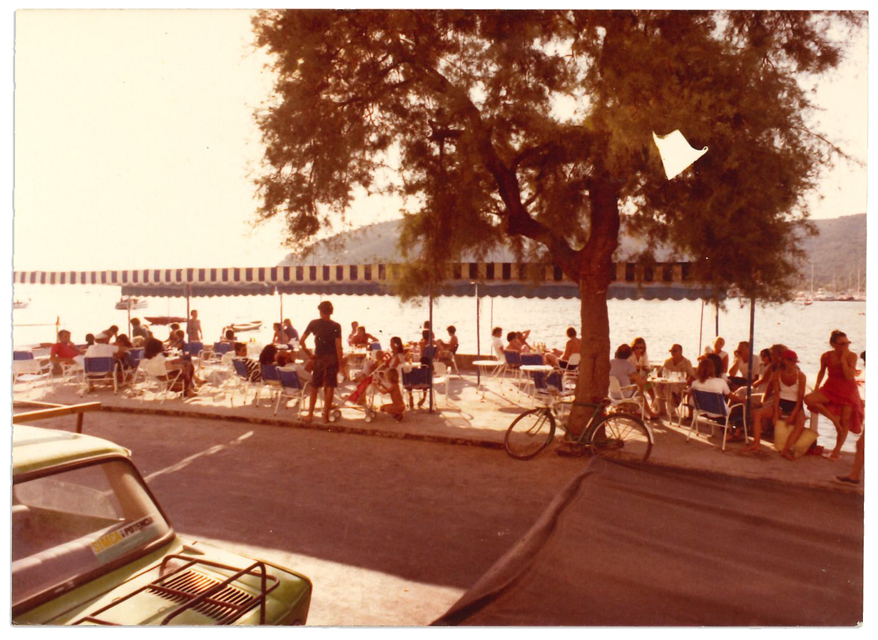 Una escena en una terraza junto al agua con muchas personas sentadas en mesas y sillas, algunos conversando y otros disfrutando del paisaje, bajo una sombrilla y un árbol grande, con un bote en el agua y un muelle en el fondo.