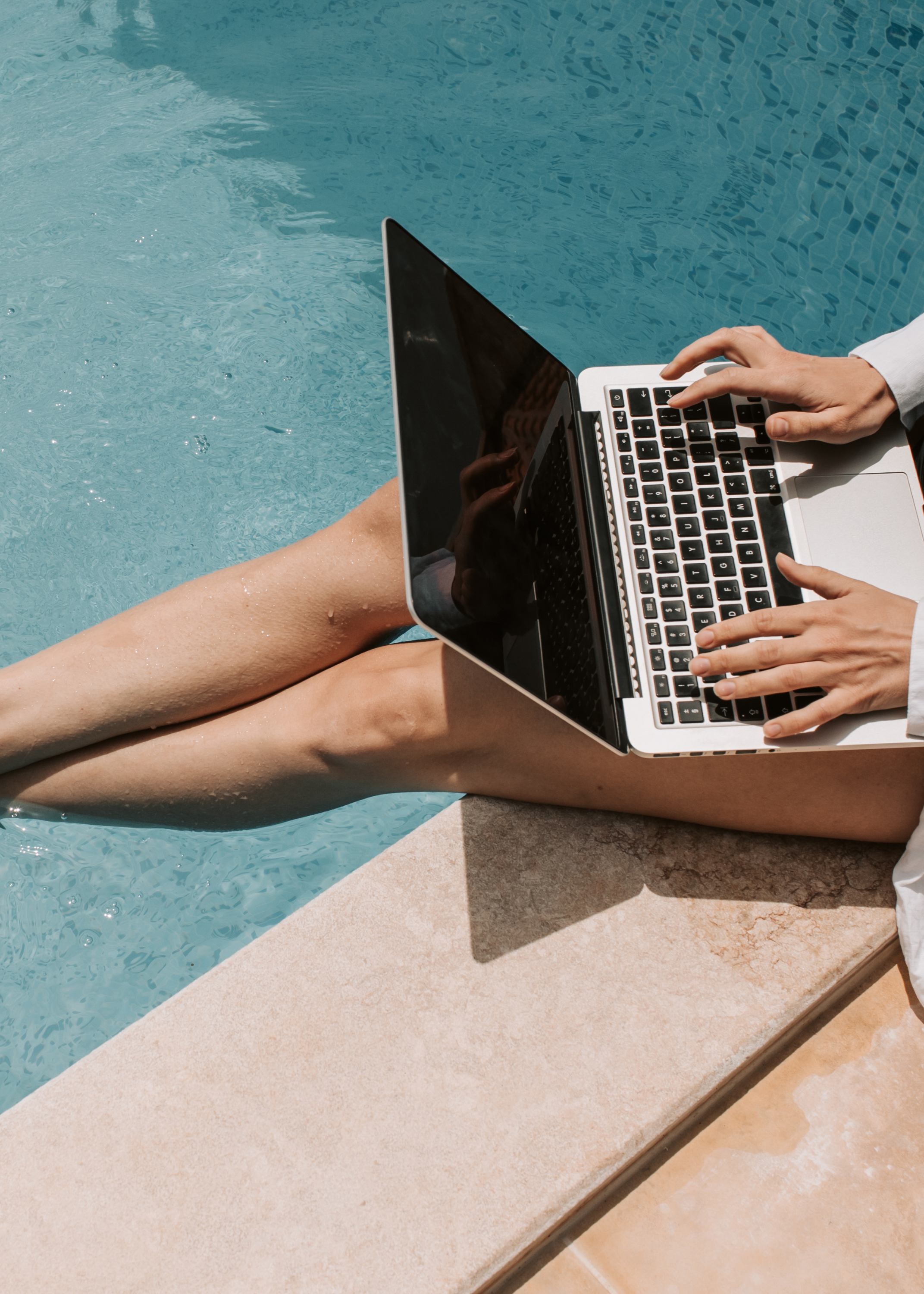 Person sitting beside a swimming pool using a laptop with water in the background