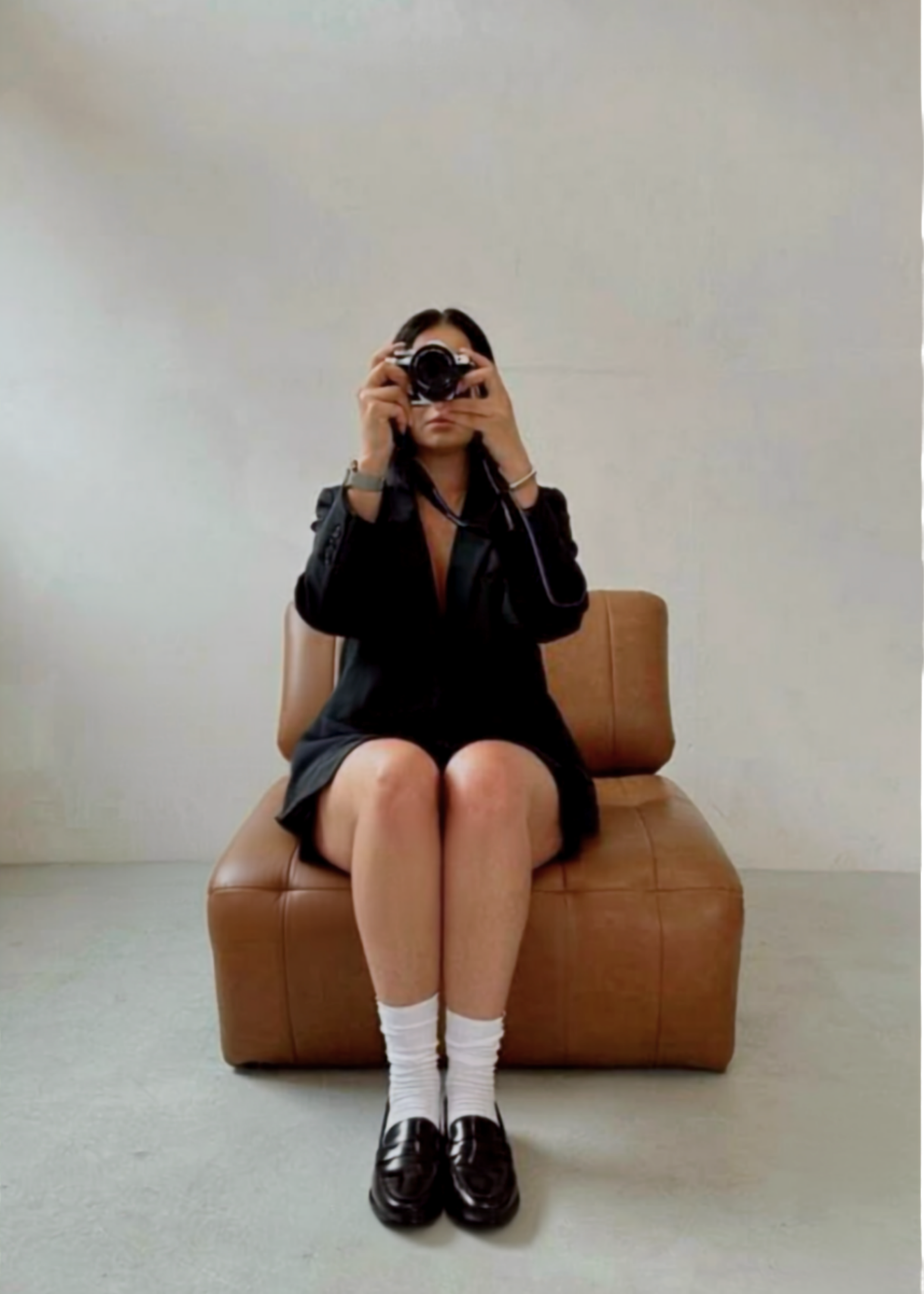 A young person sitting on a brown leather bench, holding a camera up to their face, with their knees crossed at the ankles. They are wearing a black jacket, a black skirt, white socks, and black shoes. The background is a plain, light-colored wall.