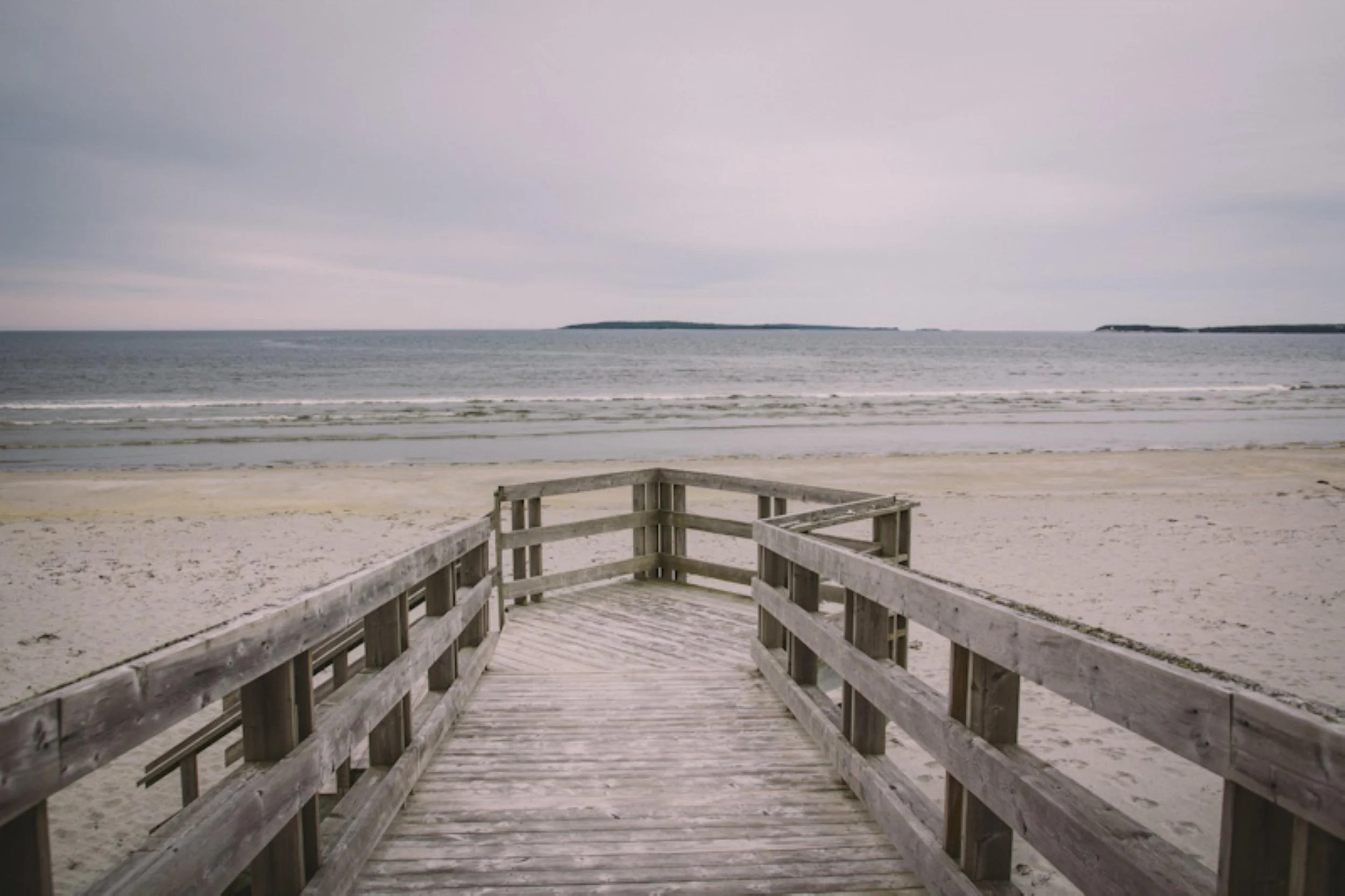 wooden walkway leading to the beach with sandy shore and ocean waves, overcast sky, distant islands or landforms in the horizon