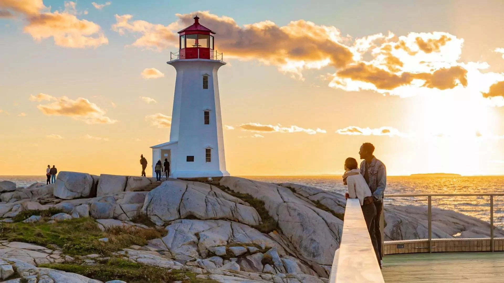 People standing on a rocky shoreline near a white lighthouse with a red top during sunset, with clouds in the sky and the ocean in the background.