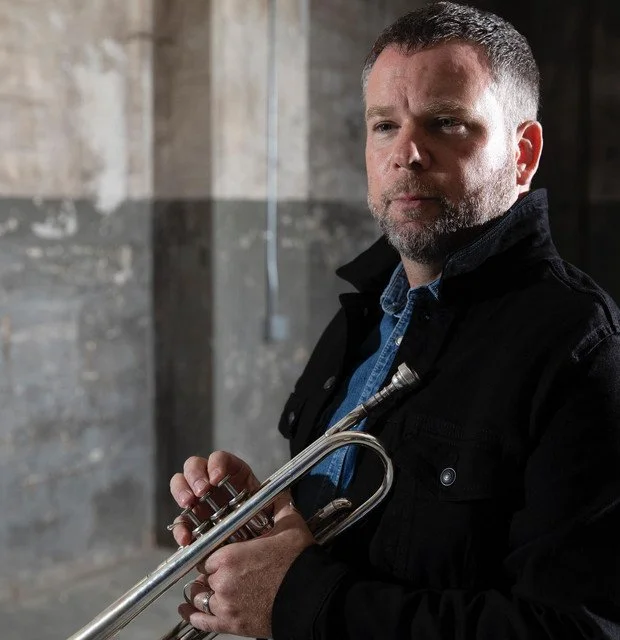 A man holding a trumpet in an industrial-style setting with exposed pipes and concrete walls.