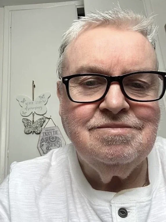 A close-up of a middle-aged man with short gray hair, glasses, and a beard, smiling slightly indoors. In the background, decorative butterfly wall art and a door with a hook are visible.