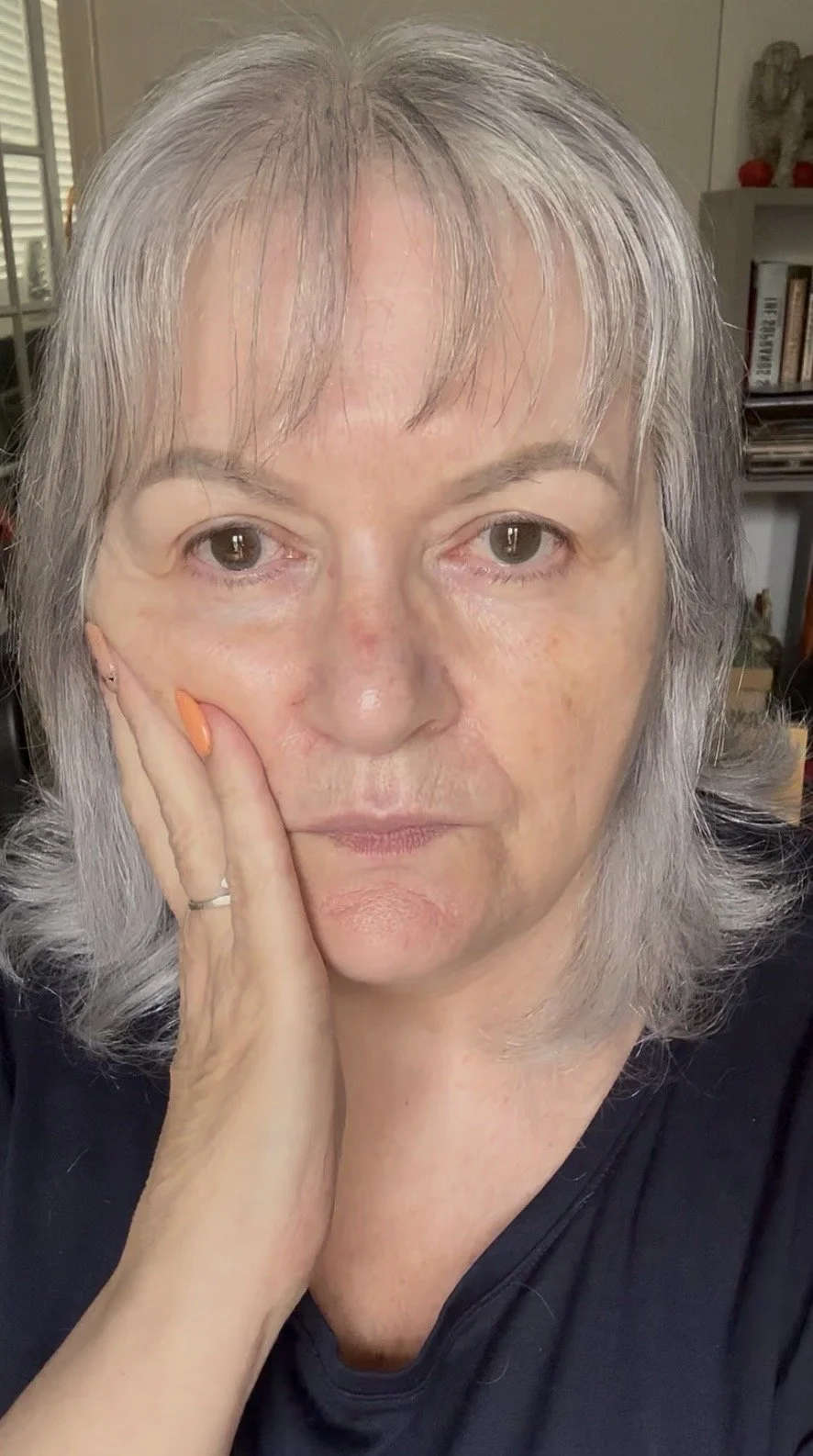 A woman with gray hair, holding her face with her left hand, wearing a black top, in a room with books and decorations in the background.