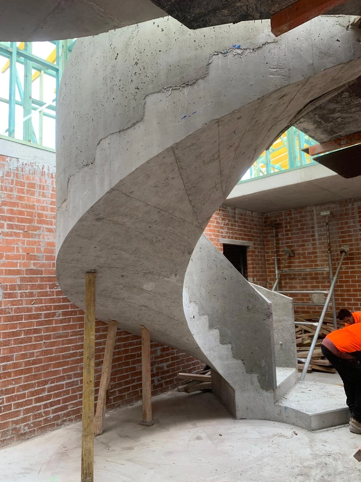 Concrete spiral staircase under construction inside a building with brick walls and construction workers.