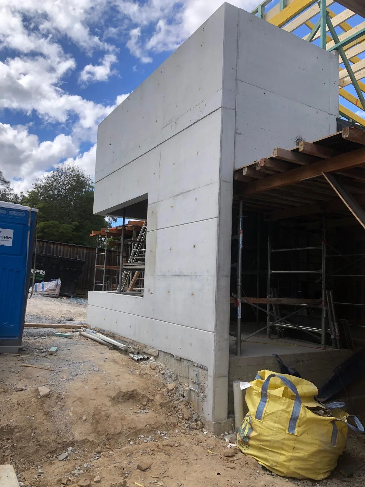 Construction site with a concrete wall and scaffolding, blue sky with clouds, power tools, and construction materials on the ground.