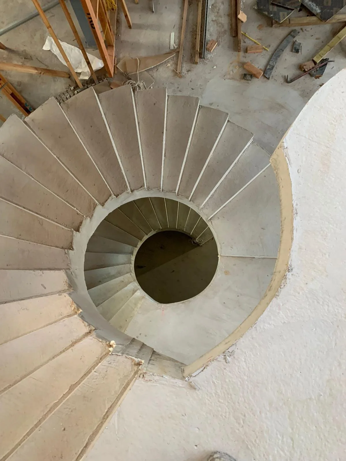 Looking down at a spiral staircase under construction, showing unfinished steps and surrounding construction area with tools and construction materials.