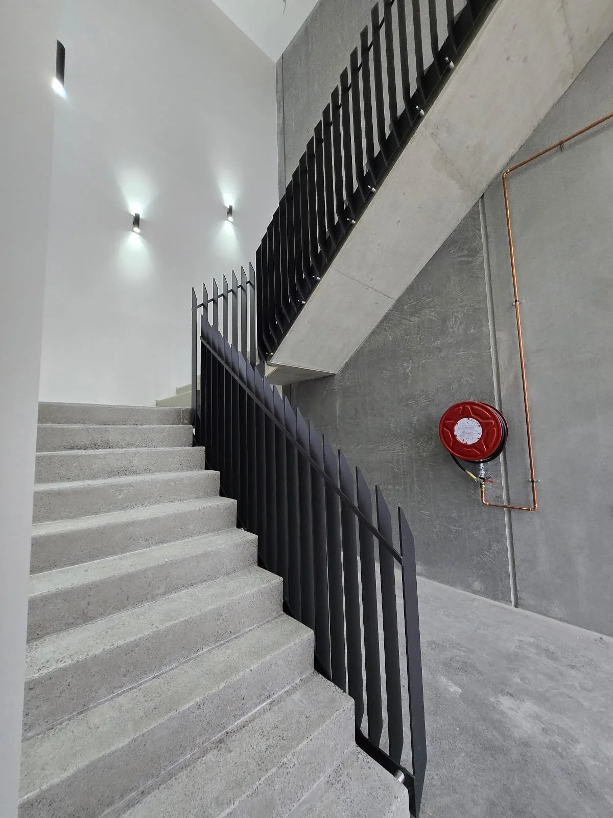Concrete staircase with black metal railing, gray walls, and a red fire hose reel on the wall.