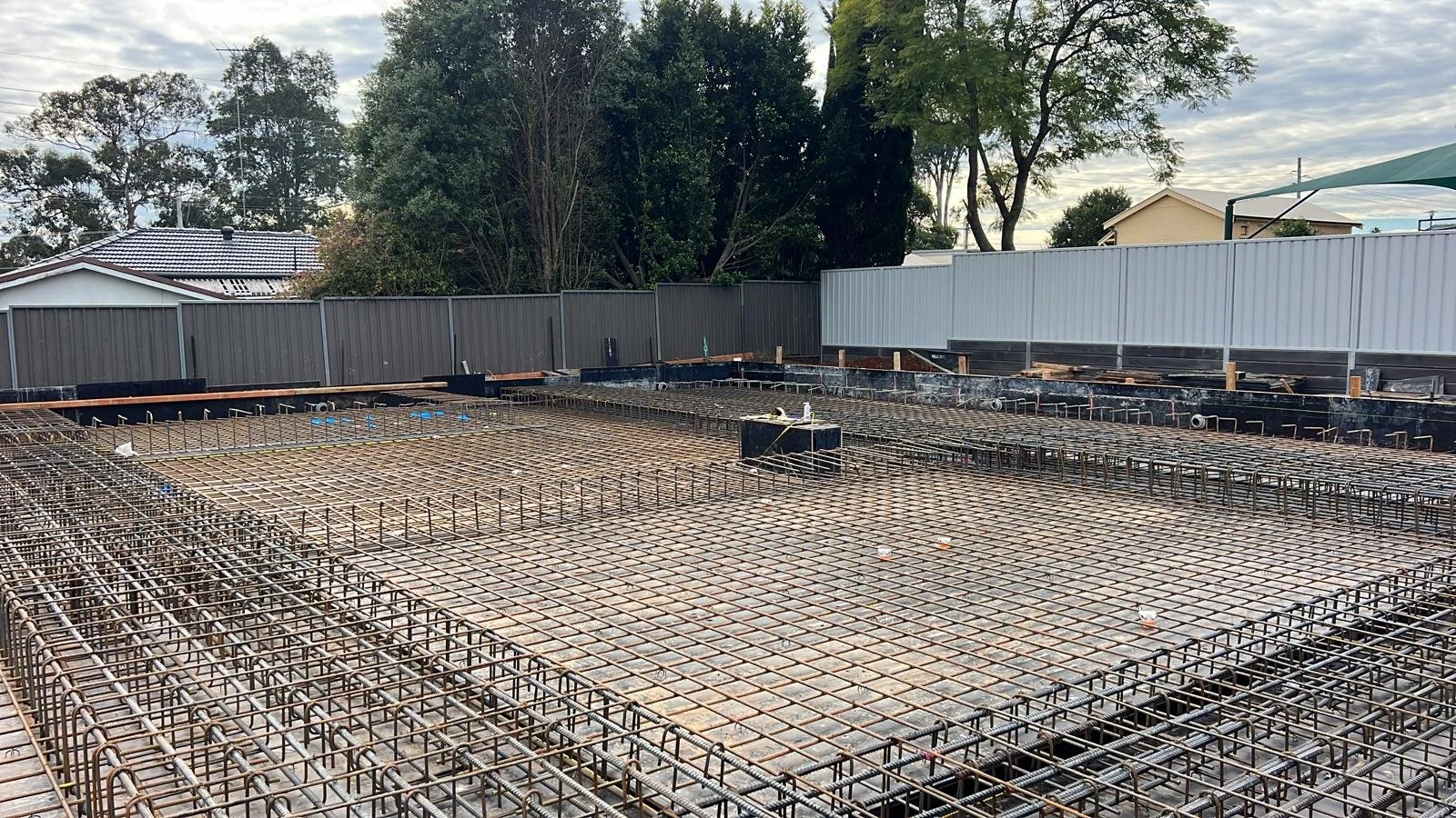 Construction site with steel rebar framework for concrete slab, surrounded by metal fencing, with trees and houses in the background.