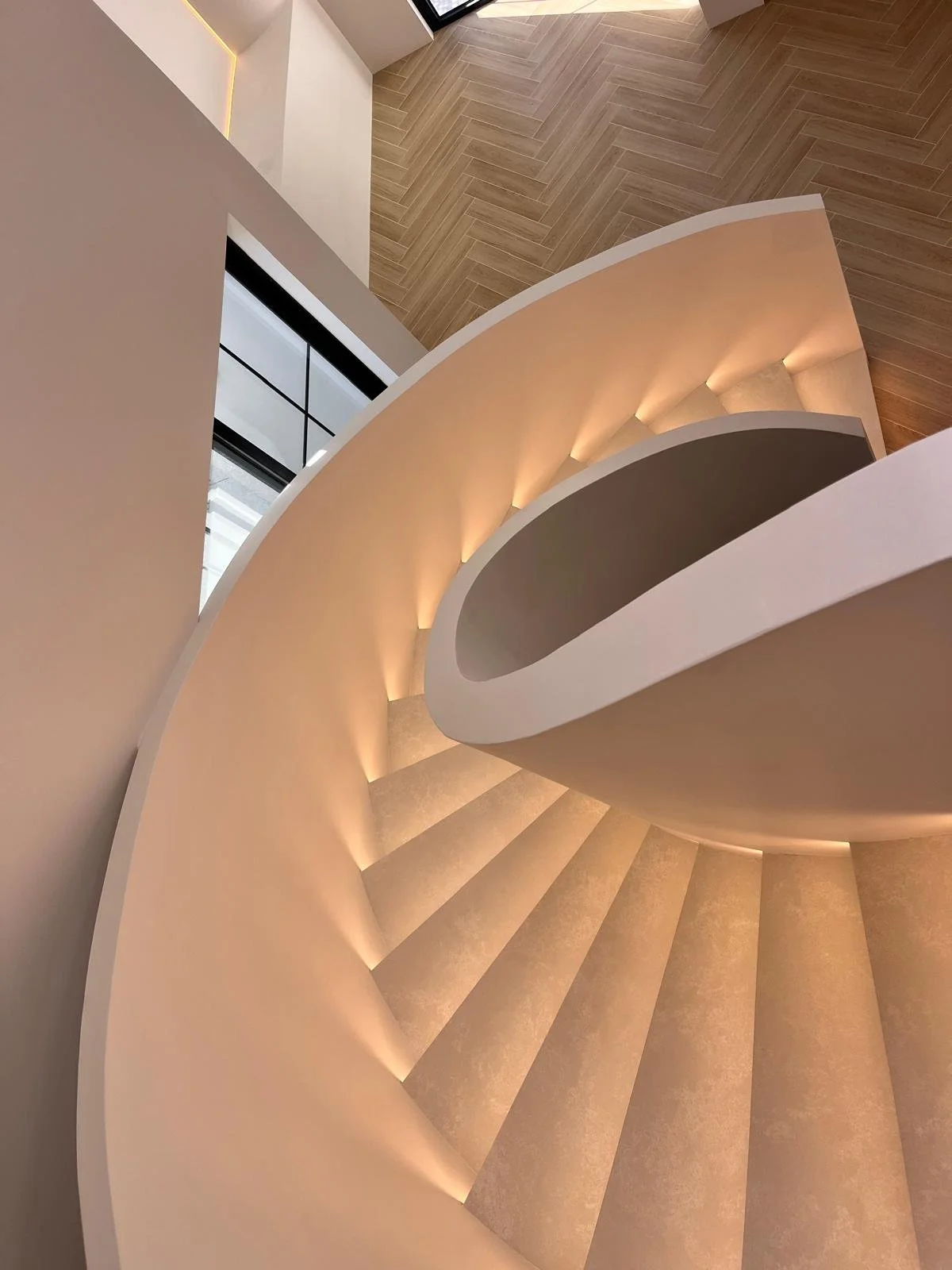 Interior view of a curved, modern staircase with warm lighting, white walls, wooden flooring, and large windows.