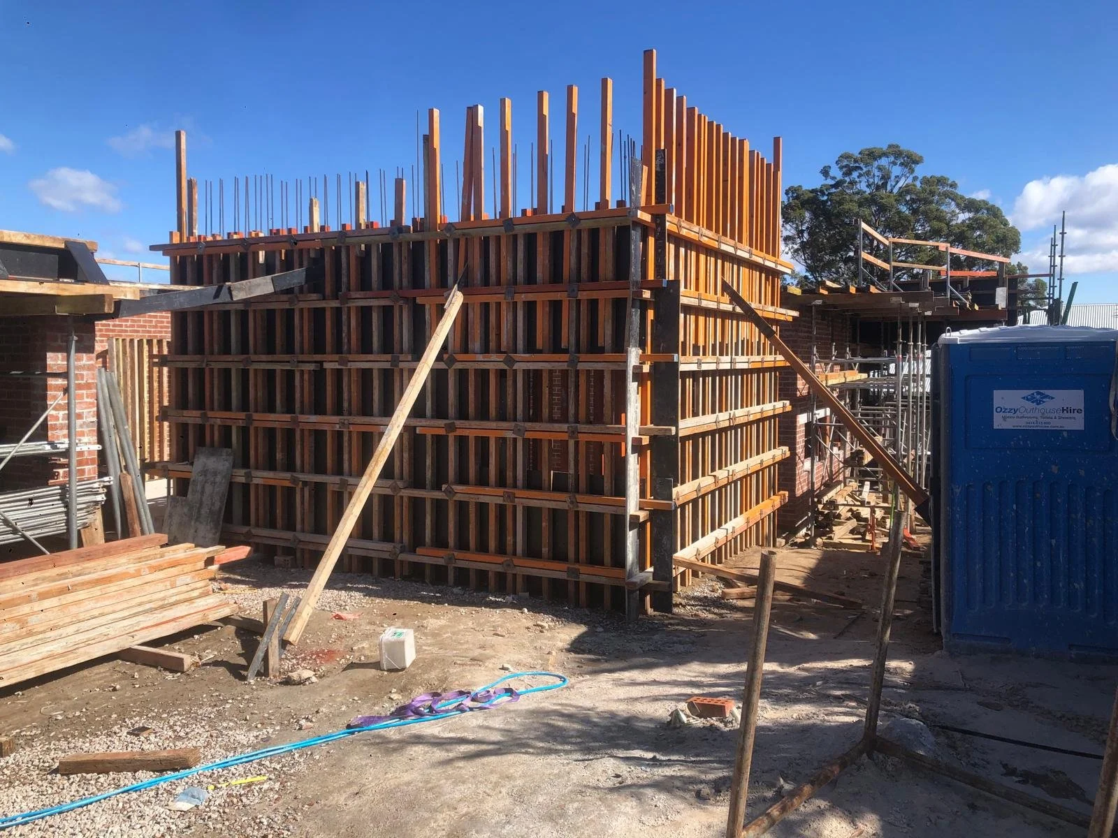 Construction site with wooden formwork and scaffolding for a building's concrete wall, blue portable toilet to the right, and building materials scattered on ground.