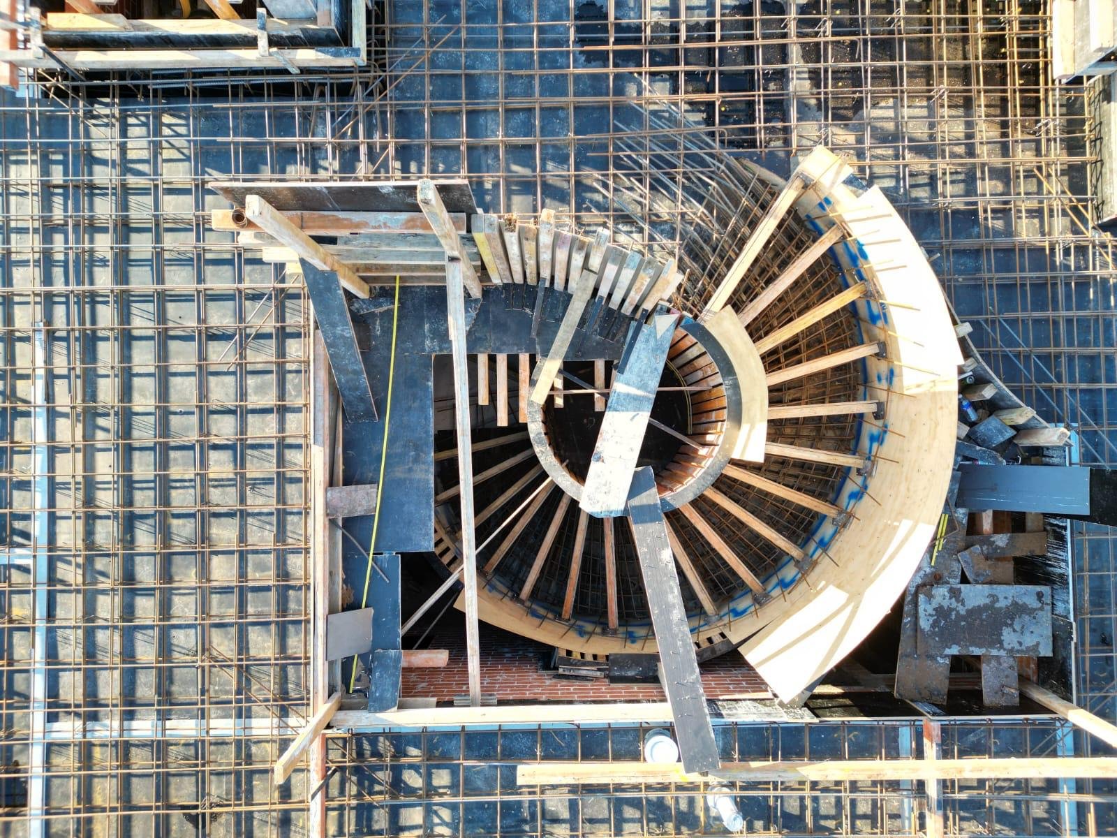 Construction site view from above showing a spiral staircase under construction, surrounded by metal scaffolding and rebar.