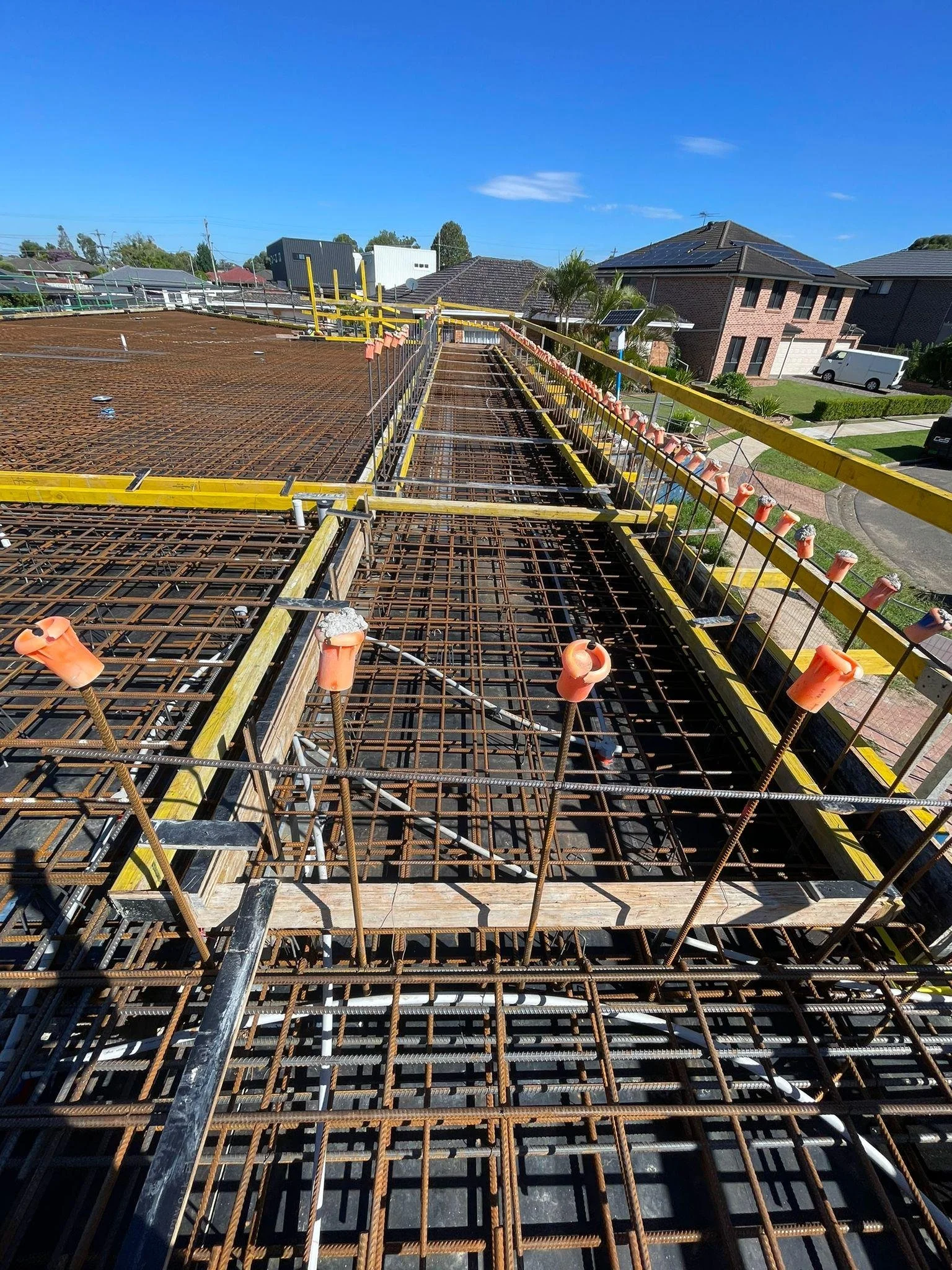Construction site with rebar and wooden formwork ready for concrete pouring, safety fencing along the edge, and residential houses in the background under a blue sky.