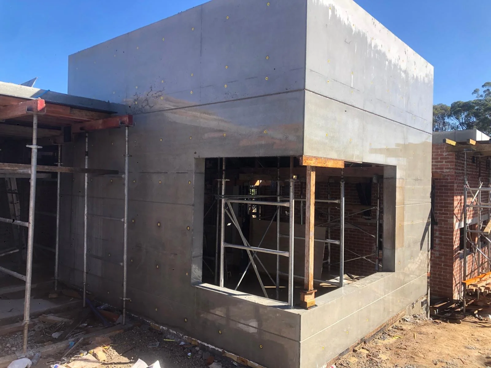 Construction site showing a building with concrete walls, metal scaffolding, and a partial brick wall on the right, under a clear blue sky.
