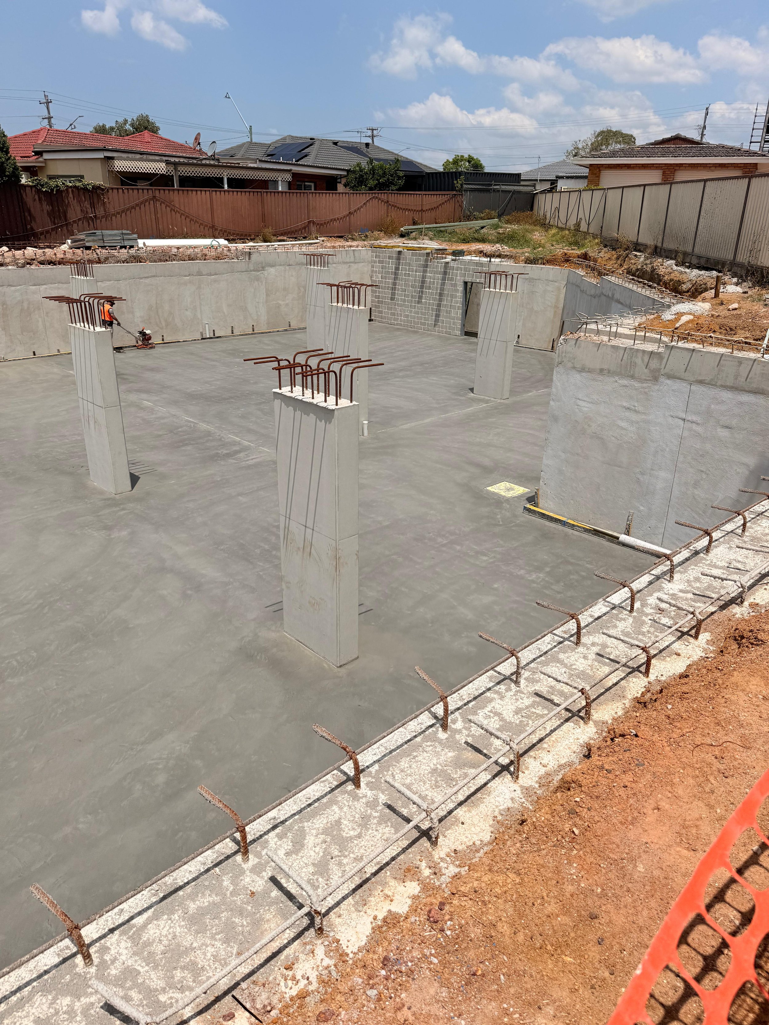 Construction site showing a concrete foundation with metal rebar protruding, surrounded by residential houses with fences under a partly cloudy sky.