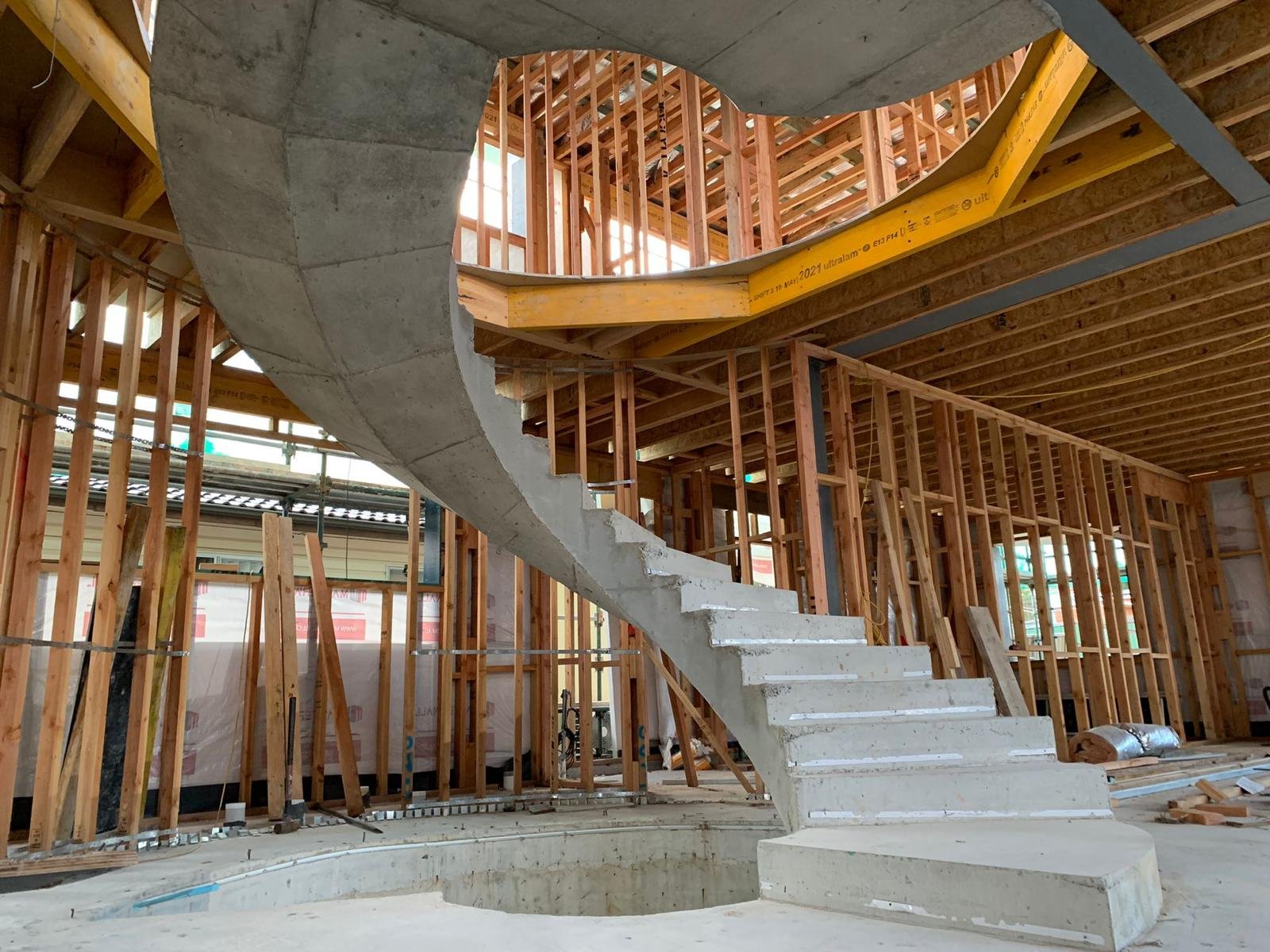 Interior view of a building under construction, featuring a curved concrete staircase with steps leading up, exposed wooden framing, and partially finished walls and ceiling.