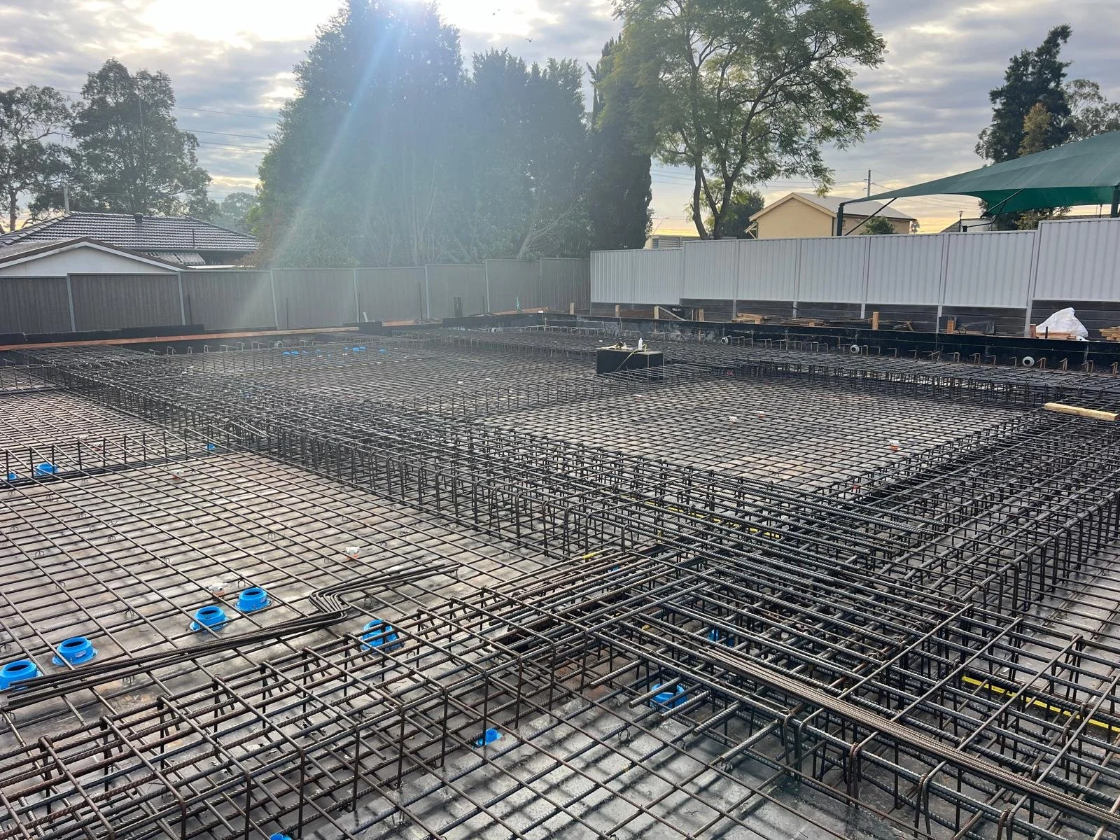 Construction site with steel rebar arranged for a concrete slab, surrounded by fences and nearby houses and trees.