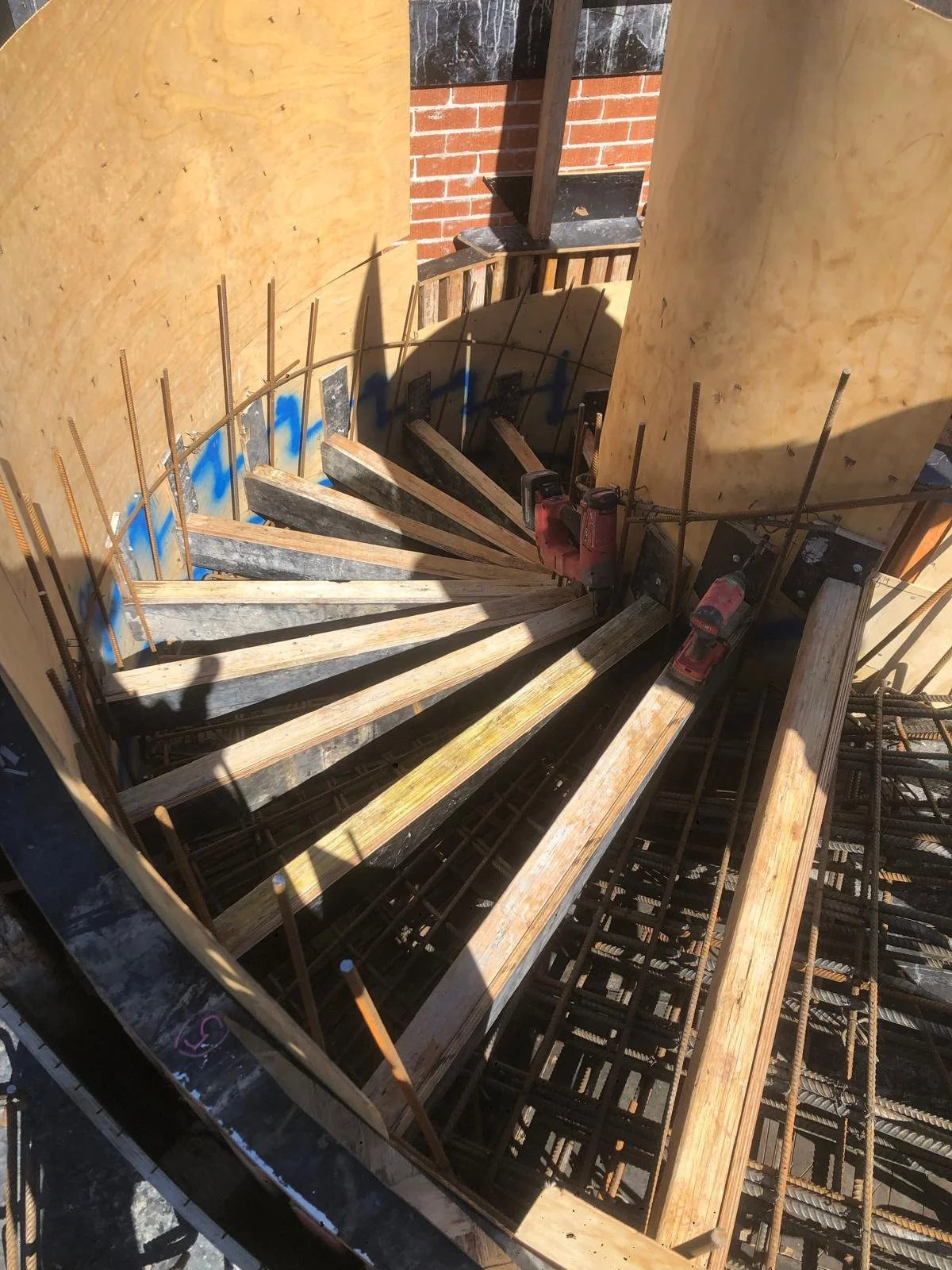Construction site showing a spiral staircase under construction with wooden forms, rebar, and power tools, near a brick wall.