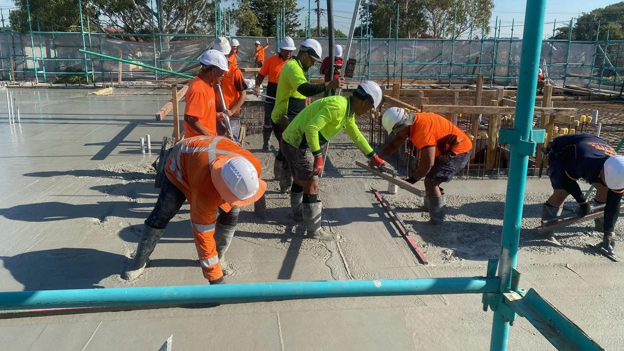 Construction workers wearing safety gear, including helmets and high-visibility clothing, are pouring and leveling concrete on a building site.